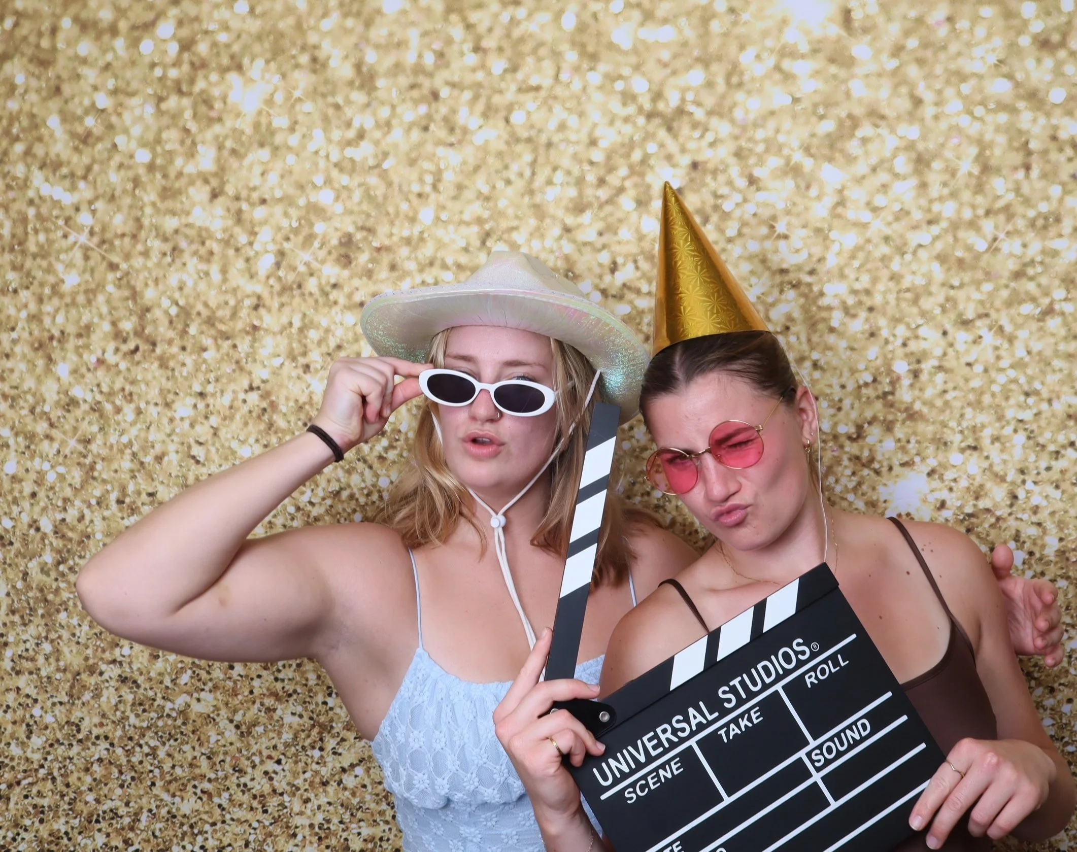 Two young women wearing party hats and sunglasses, holding a film clapperboard, with a sparkling gold background. Posing in front of a photobooth.