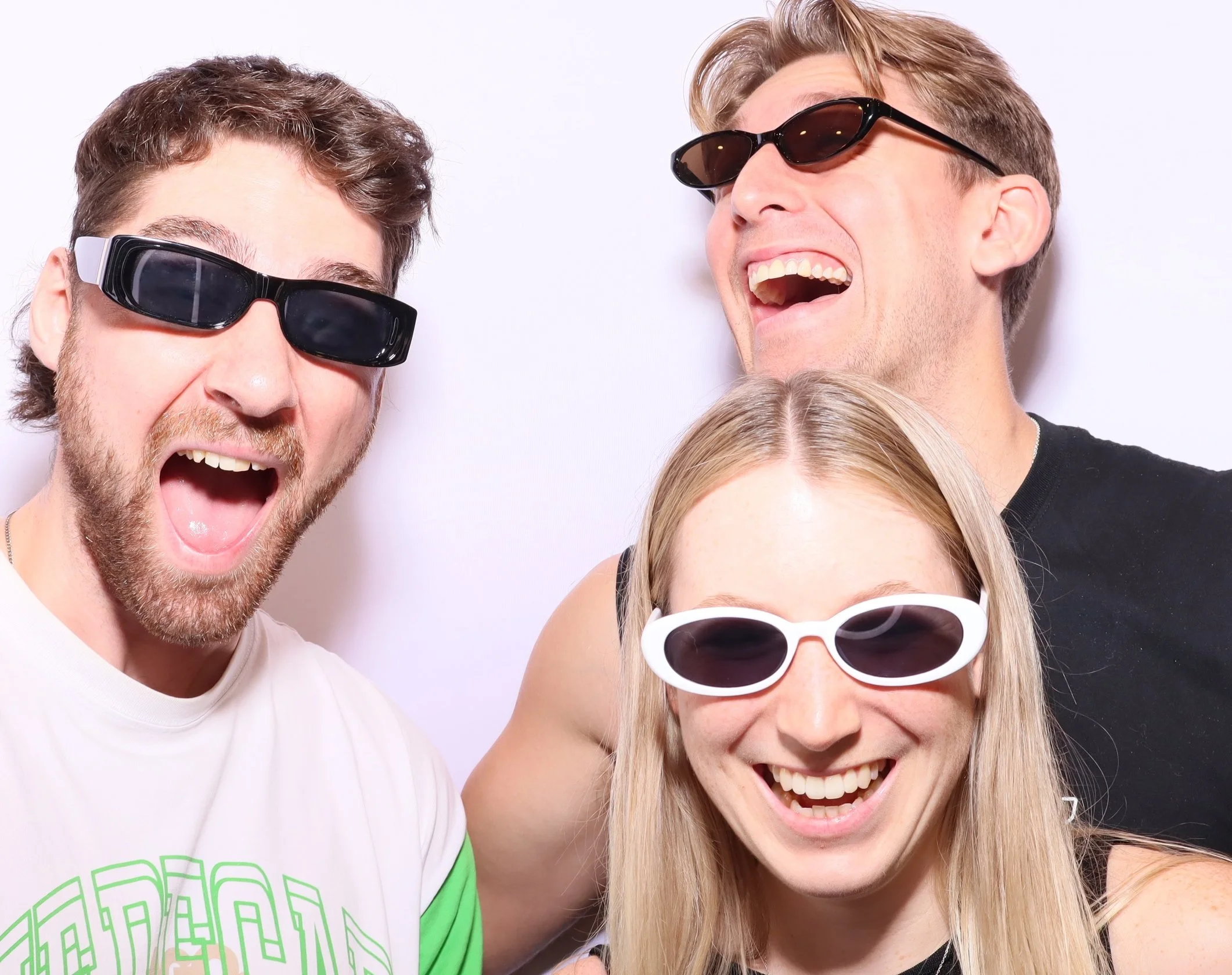 Three young adults, two men and one woman, wearing sunglasses and smiling or laughing, taking a photo together indoors against a plain white background. Posing in front of a photobooth.