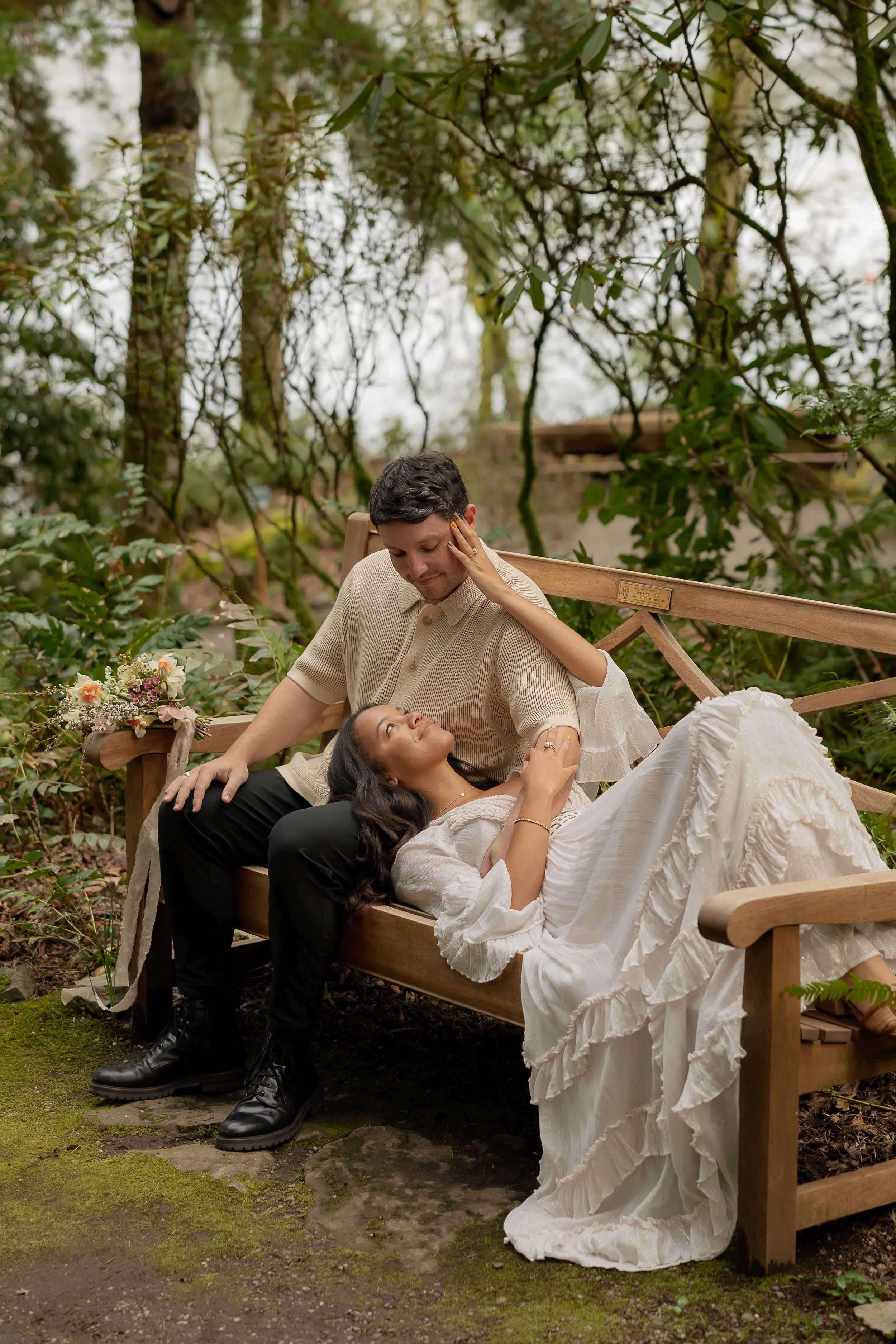 A man and woman sharing an intimate moment on a wooden bench outdoors, surrounded by trees and greenery. The woman is lying down with her head on the man's lap, looking up at him, while he gently touches her face.