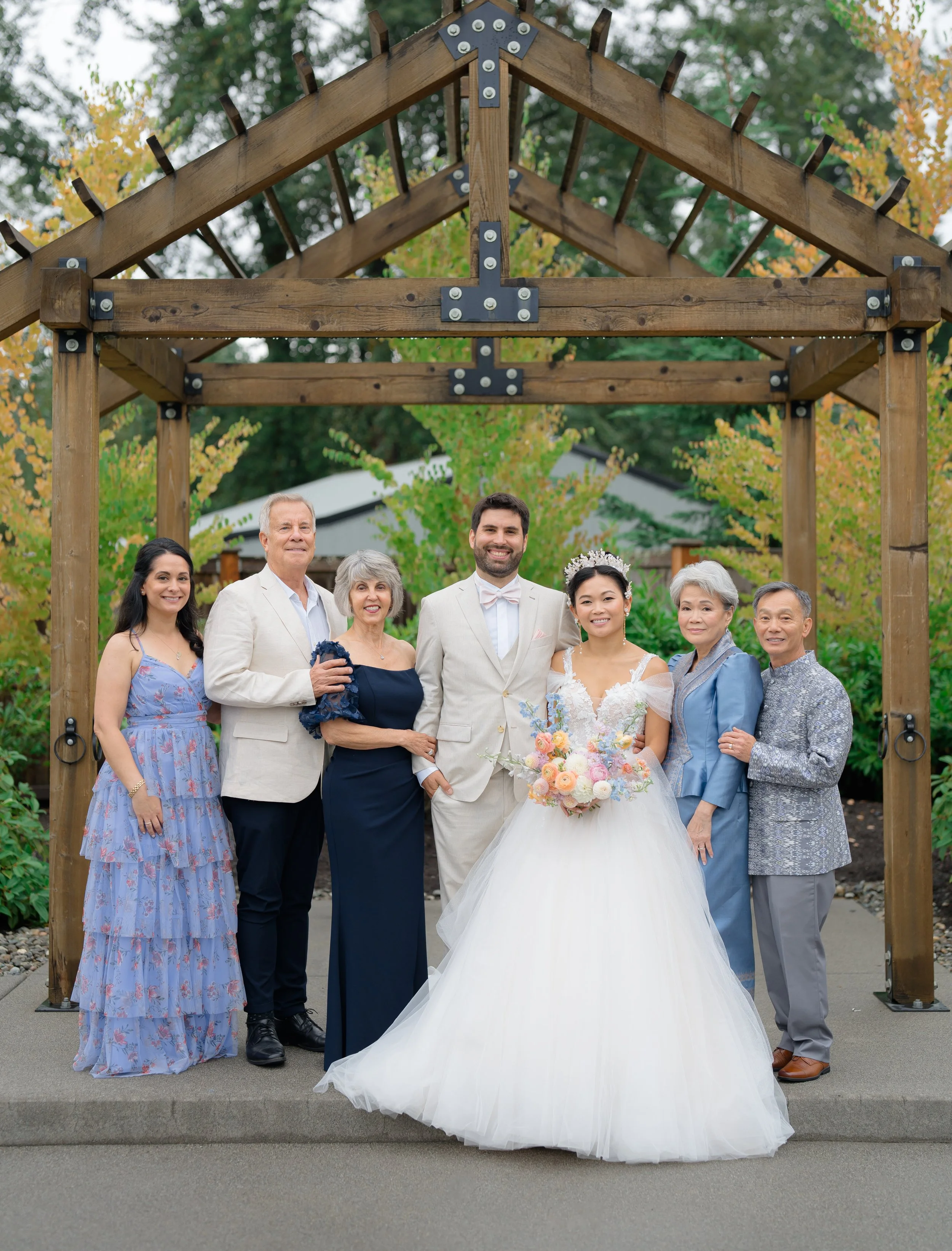 A group of seven people, including a bride in a white gown holding a bouquet, stand smiling under a wooden arbor. They are dressed in formal attire, and the setting is an outdoor venue with trees in the background.