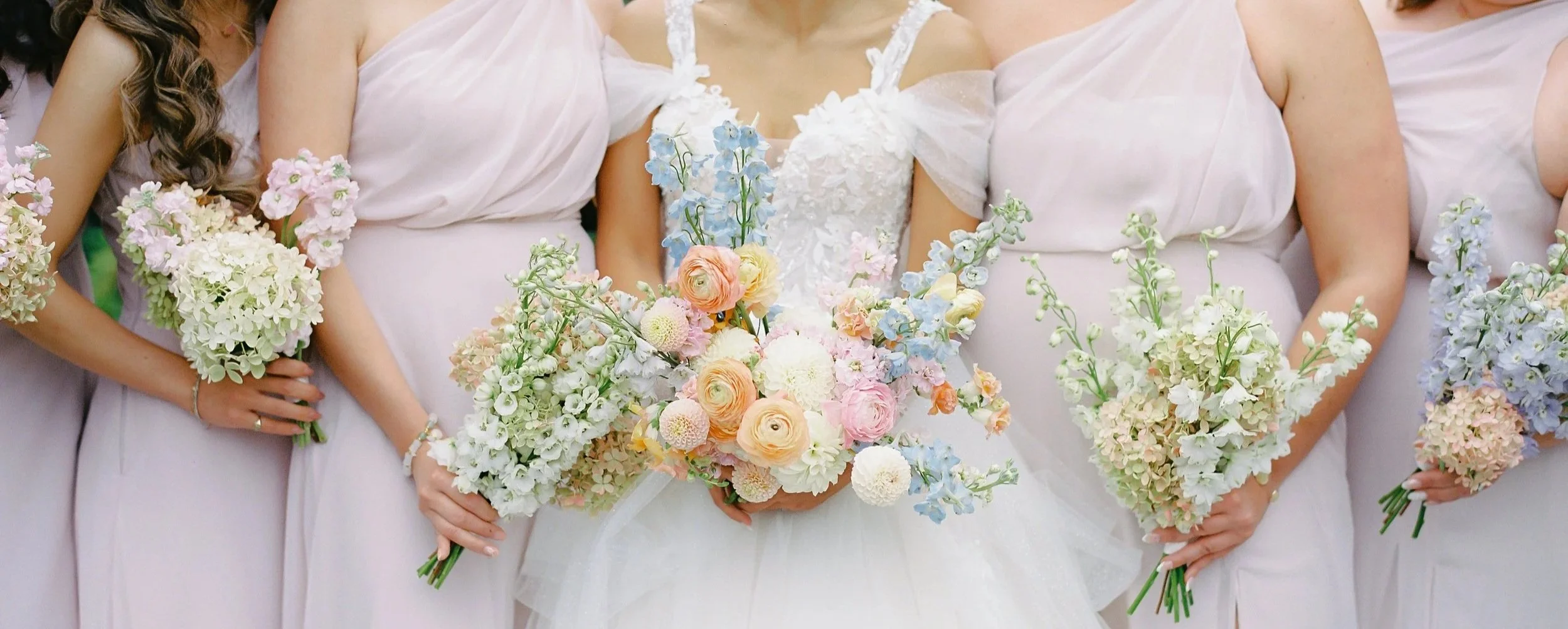 Bride and bridesmaids holding bouquets of pastel-colored flowers, standing together in wedding dresses.