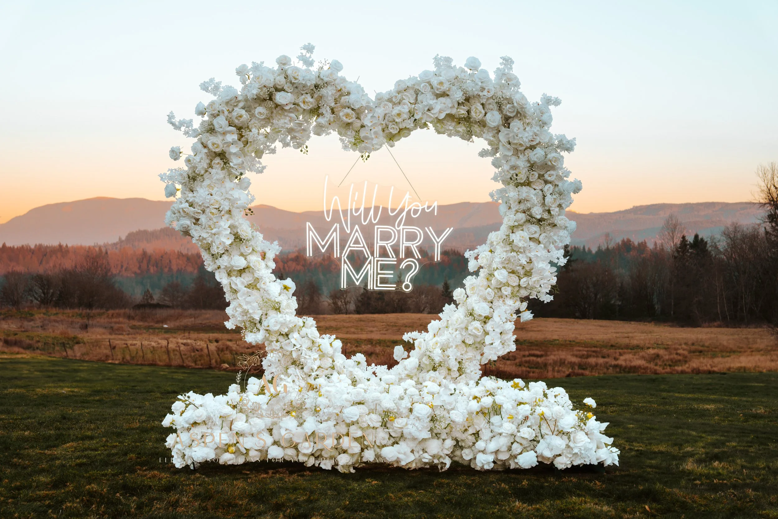 A large heart-shaped floral arrangement made of white flowers outdoors on a grassy field, with a sunset sky and mountains in the background. Text overlay asks, 'Will you MARRY ME?'.