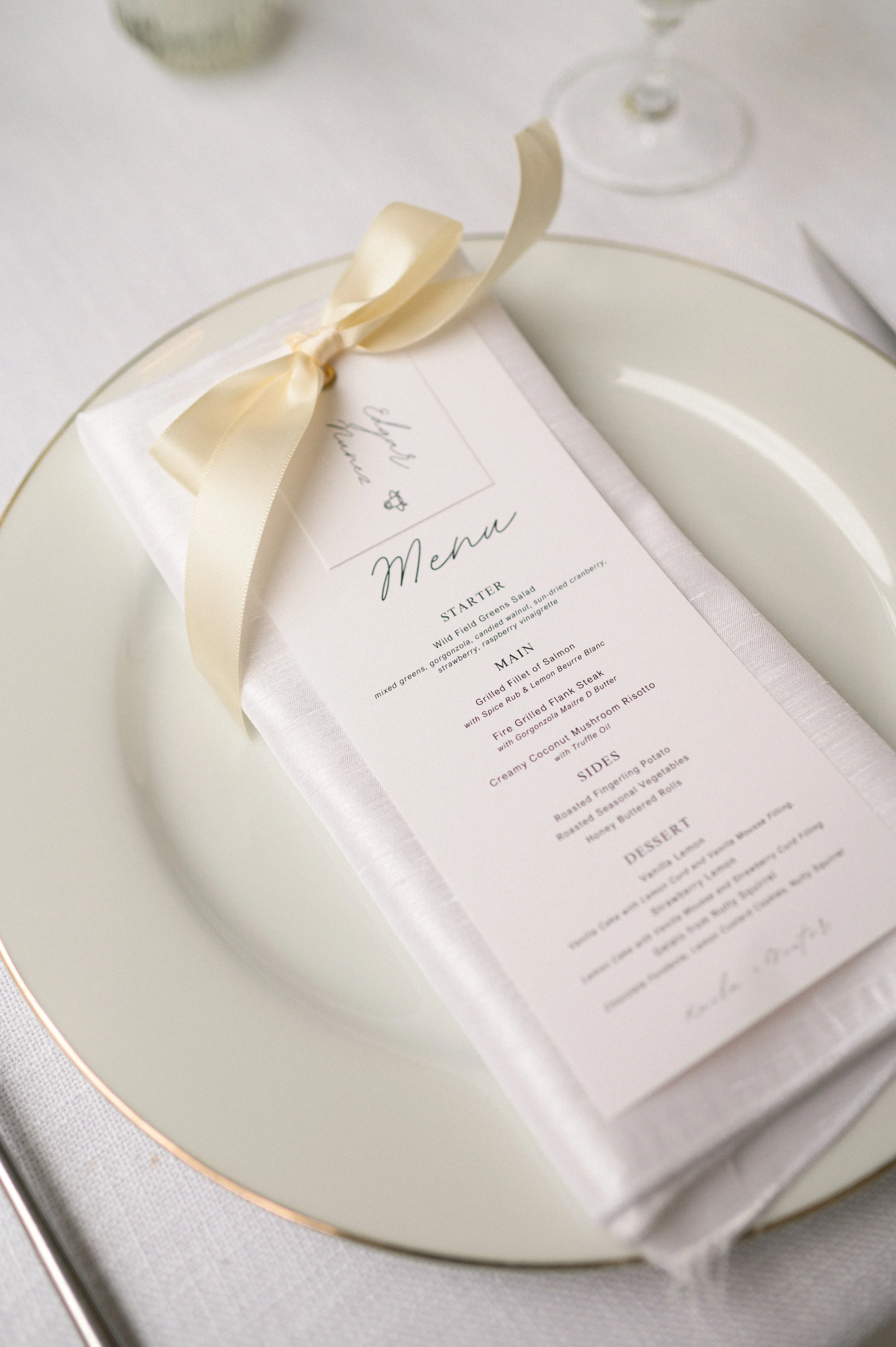 Wedding menu card with a pink flower, a blue flower, and a purple flower surrounding it. The card has elegant text listing a three-course meal and a white ribbon tied at the top. There is also a place card with the name 'Mayla' and a white tag with a gold tassel.