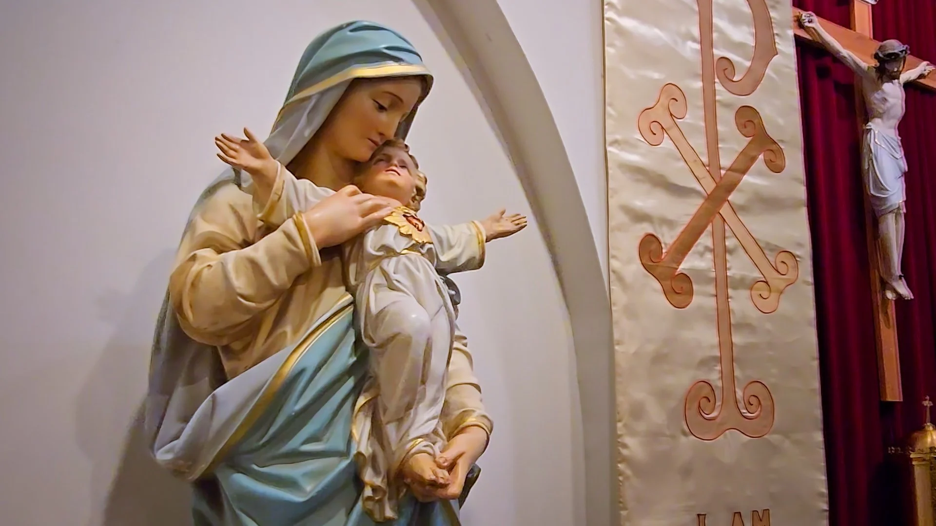 Statue of the Queen of the Daughters of the Immaculata holding baby Jesus, with both arms outstretched and cruciform. She dressed in beige, gold, and light blue robes, and Jesus Christ is in white with His Most Sacred Heart visible for contemplation.