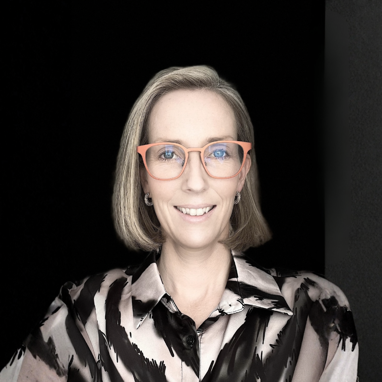 Porträtt av en kvinna med ljus hud, kort brunt hår, glasögon med rosa bågar och ett blommigt svart och vitt blus, mot en svart bakgrund.