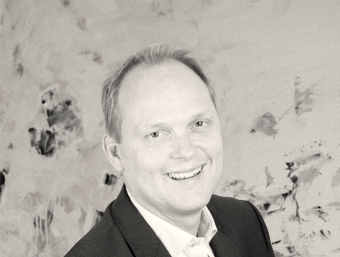 Svart-vitt porträtt av en man i formell klädsel som ler, mot en konstnärlig bakgrund.