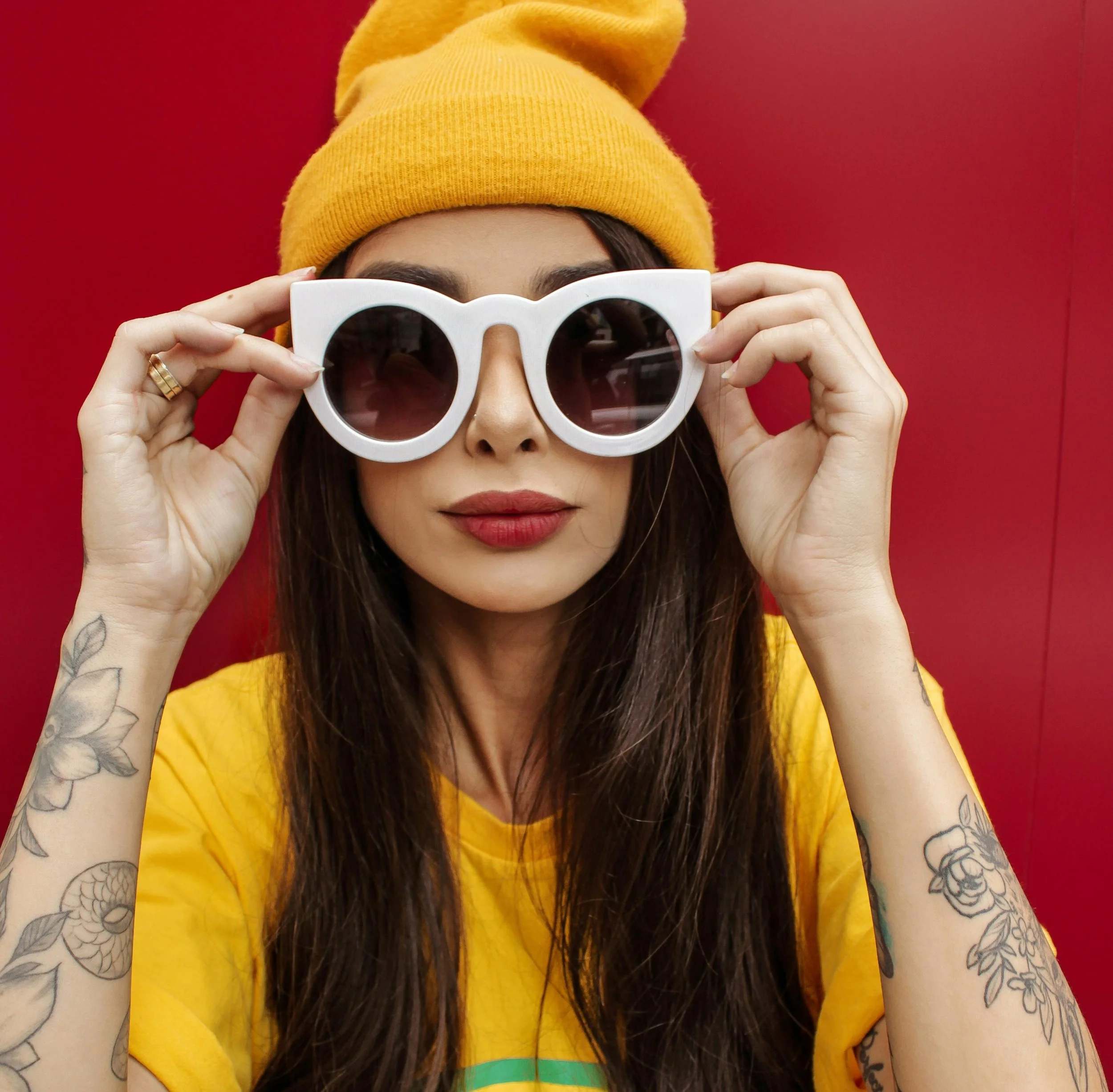 En kvinna i gul t-shirt och gul mössa med vita solglasögon mot en röd bakgrund.