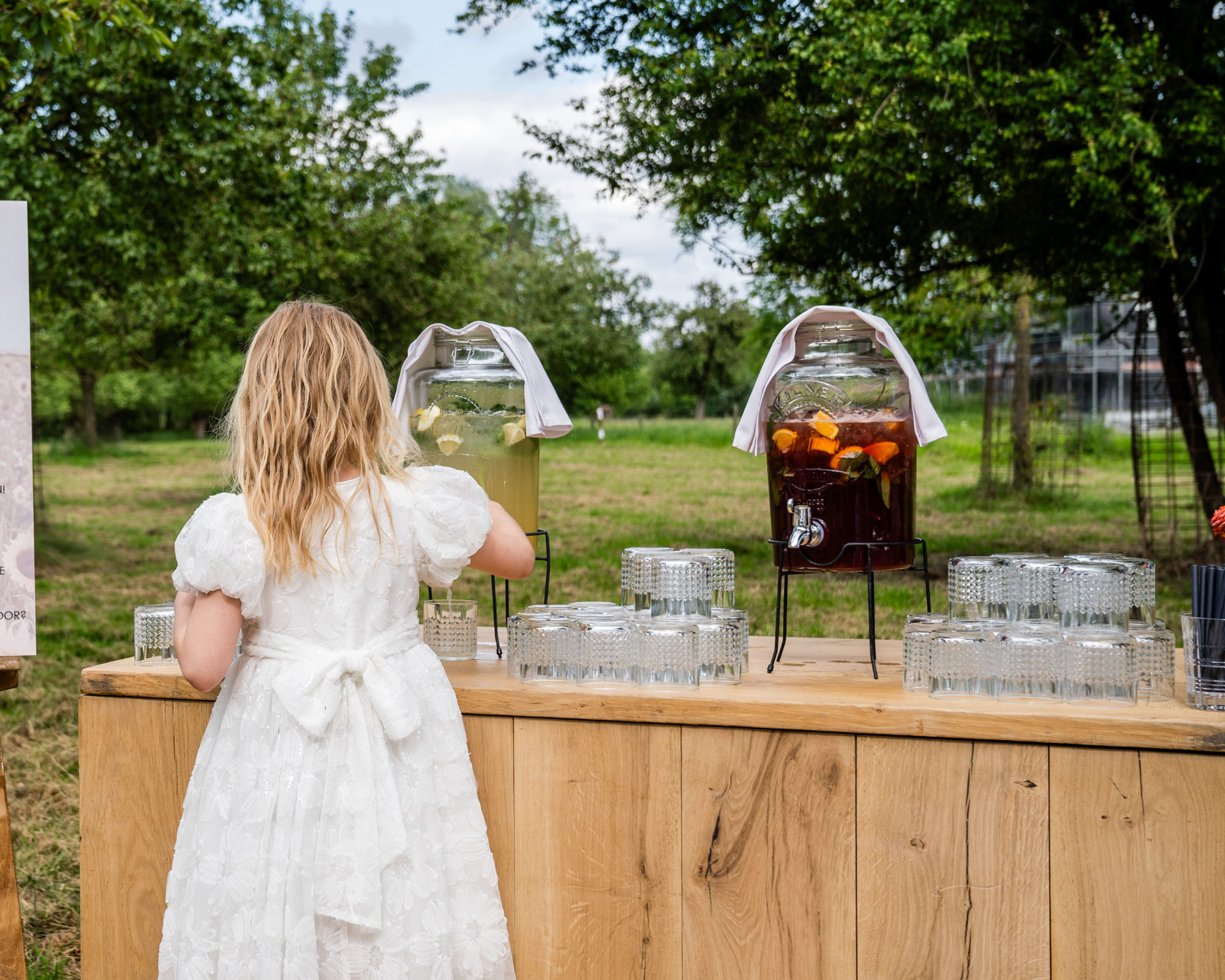 Een jong meisje in een witte jurk vult een glas met limonade bij een bar in een groene tuin.