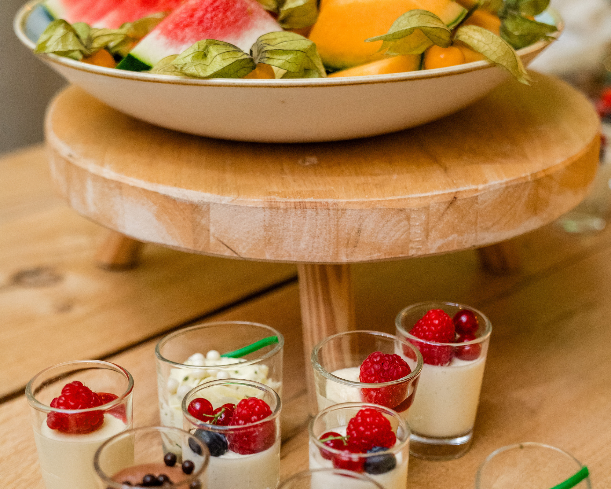 Diverse glazen met pudding of yoghurt met rood fruit zoals frambozen, kersen en bosbessen, op een houten tafel met een houten plateau en fruitschaal erboven