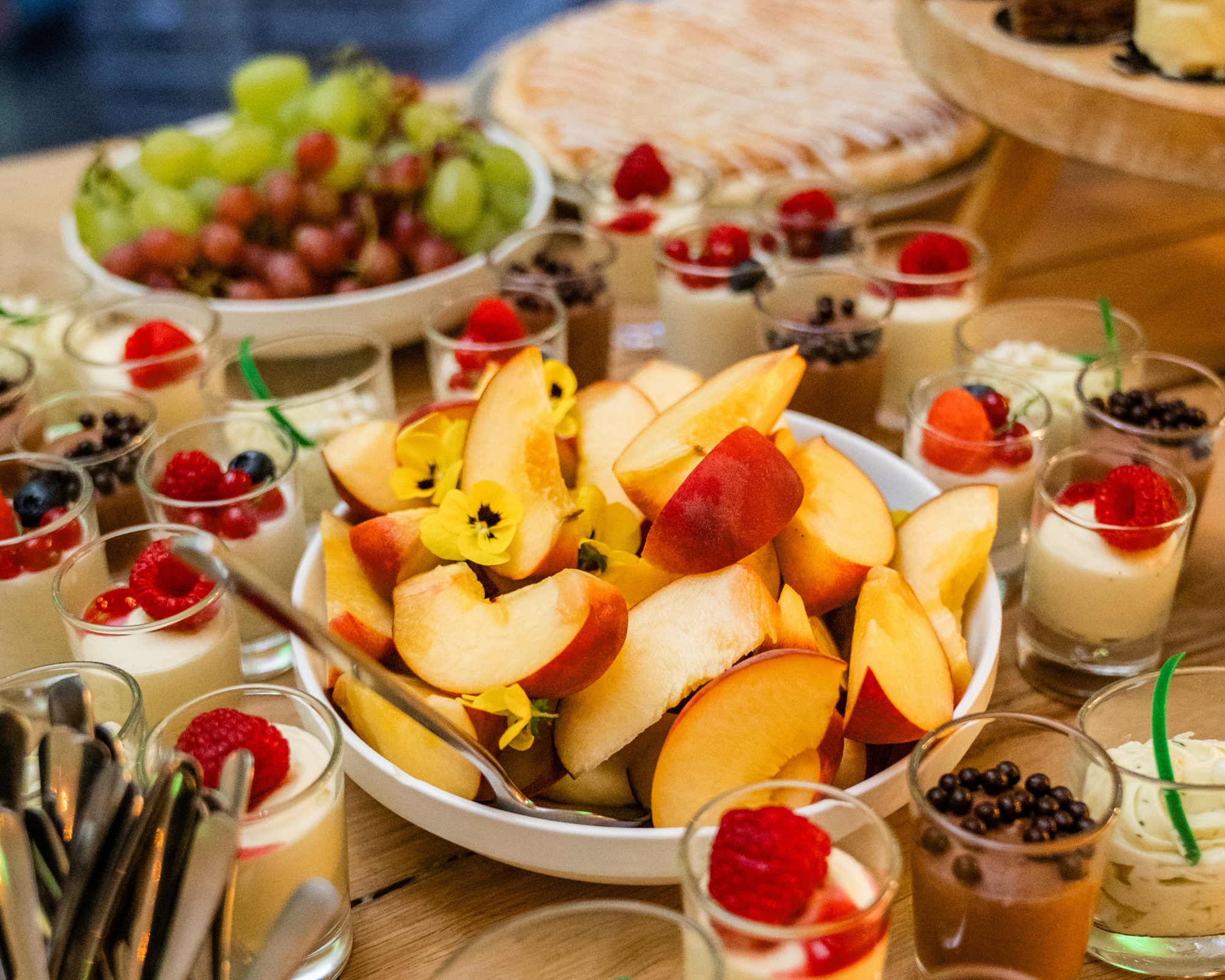 Verscheidene schaaltjes met fruit en pudding, inclusief gehakte perziken, druiven, frambozen, blauwe bessen, en creme dessert met fruit toppings op een houten tafel.