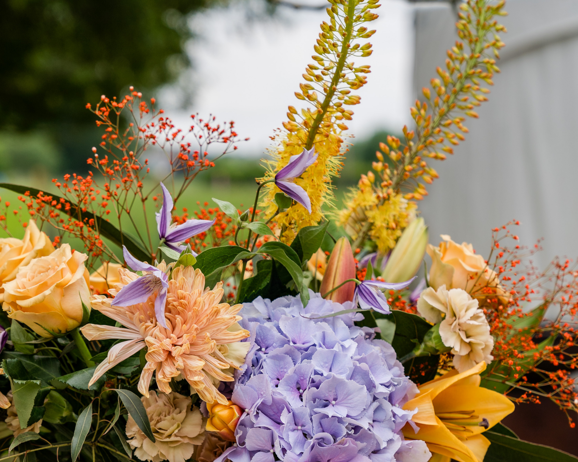 Een kleurrijk boeket met bloemen, waaronder blauwe hortensia's, lichtoranje chrysanten, paarse clematis, gele lelies, en kleine rode en gele bloemen.
