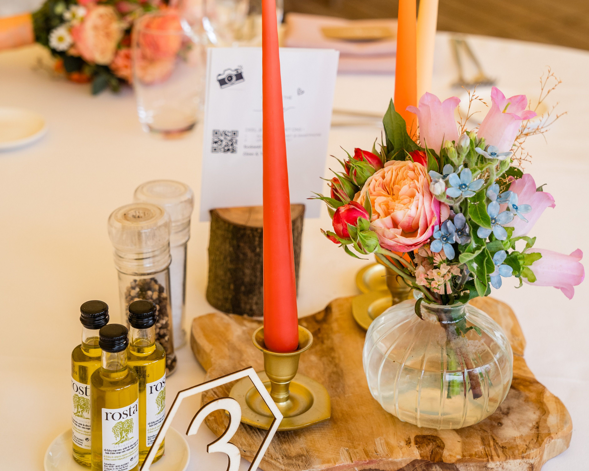 Bloemstuk met roze en blauwe bloemen in een glazen vaas op een houten bord, met twee rode kaarsen en peper- en olieflessen op een tafel. Tafeldecoratie.