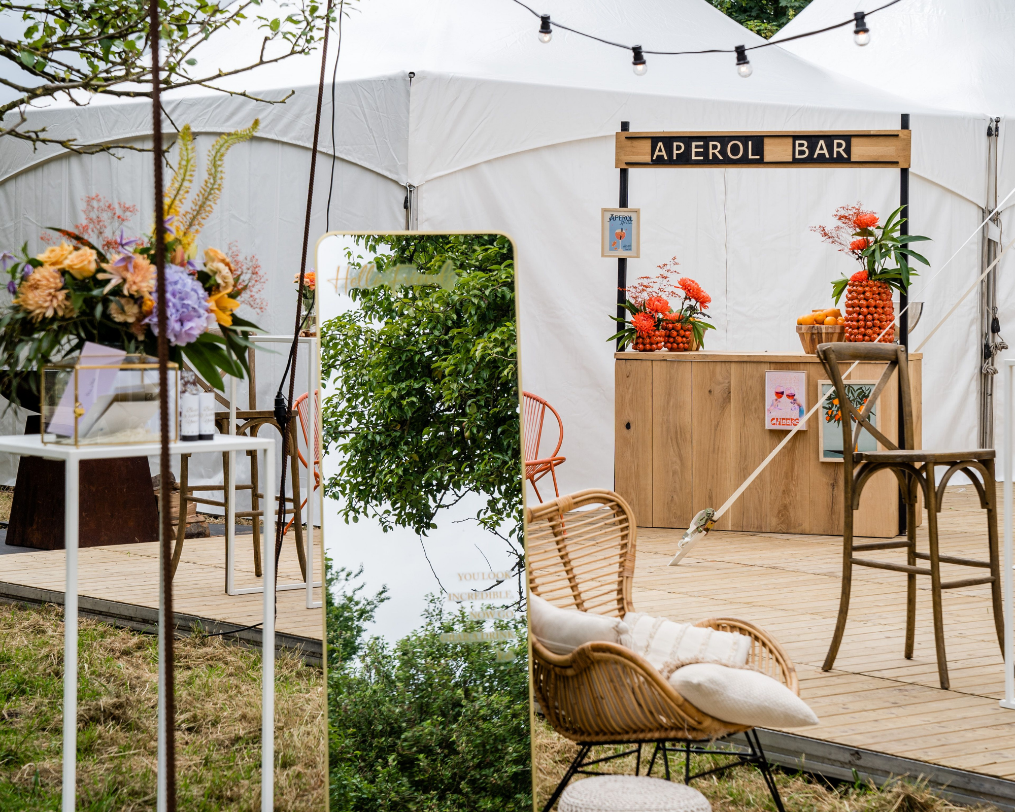 Een gezellige buitenbar met een houten bar en bloemendecoraties, onder een witte tent. Er is een grote spiegel en zitplaatsen met kussens, naast een romantisch festoon van lichtslingers.