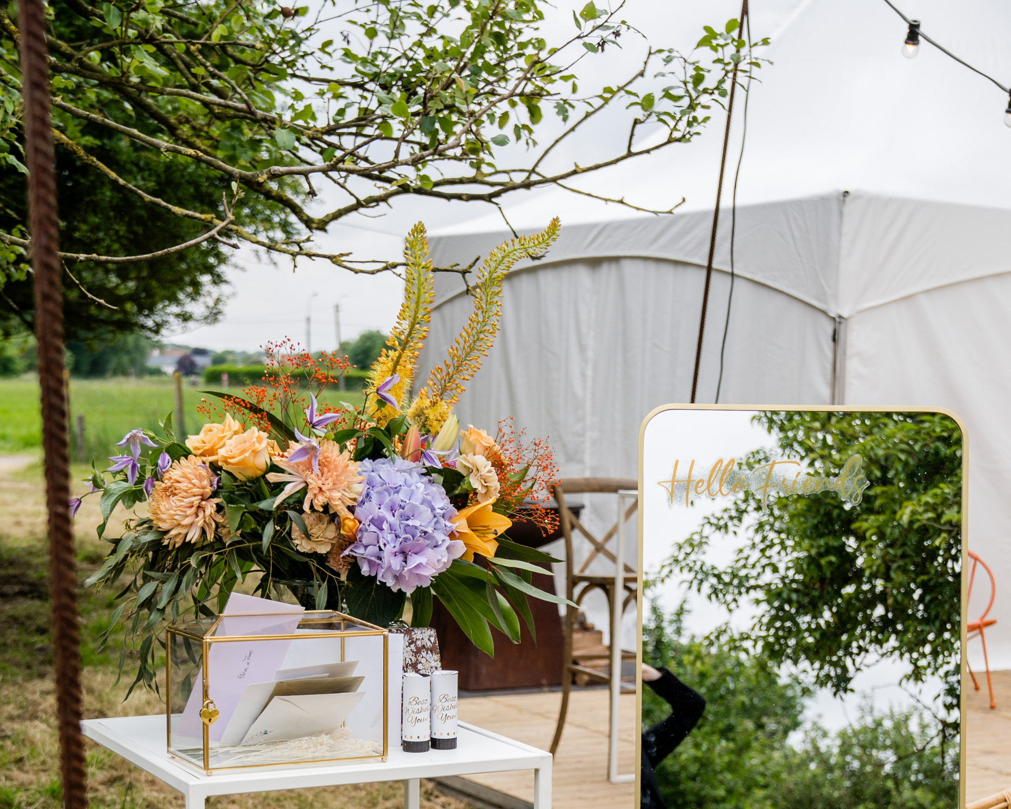 Een bloemstuk met diverse kleurrijke bloemen, een spiegel met de tekst 'Hello Friends', en een witte tafel met kaarten en kleine geschenken voor een huwelijksfeest of buitenfeest.