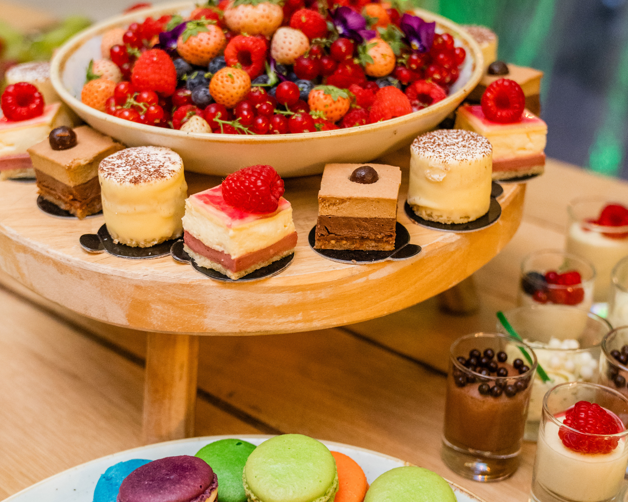 Een desserttafel met een grote schaal vol gemengd rood fruit zoals aardbeien, blauwe bessen en frambozen, omgeven door diverse kleine nagerechten en macarons. Dessertenbuffet.