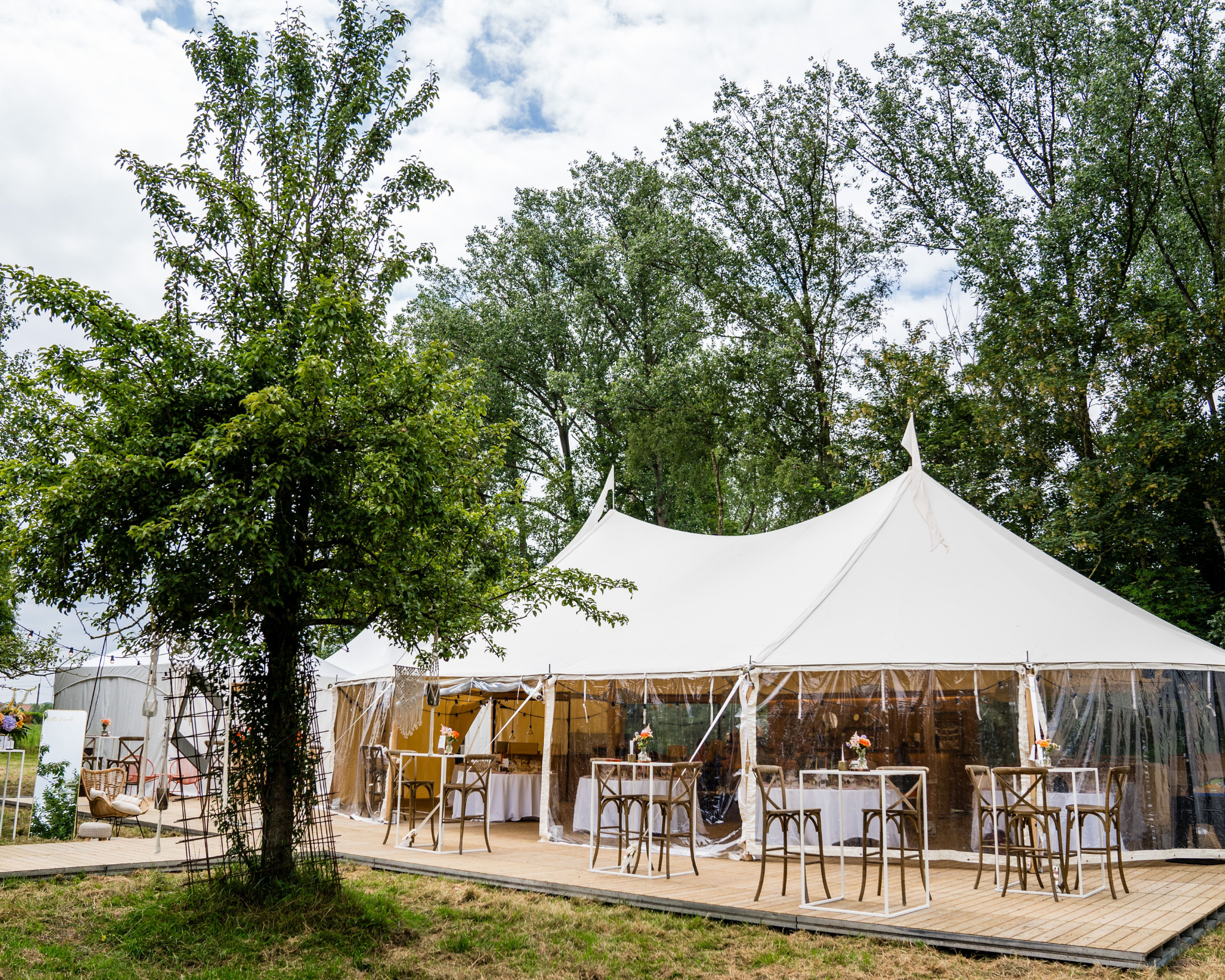 Grote witte tent met transparante zijwanden op een houten platform in een bos met groene bomen, tafels en stoelen eromheen, decoratief met bloemen.