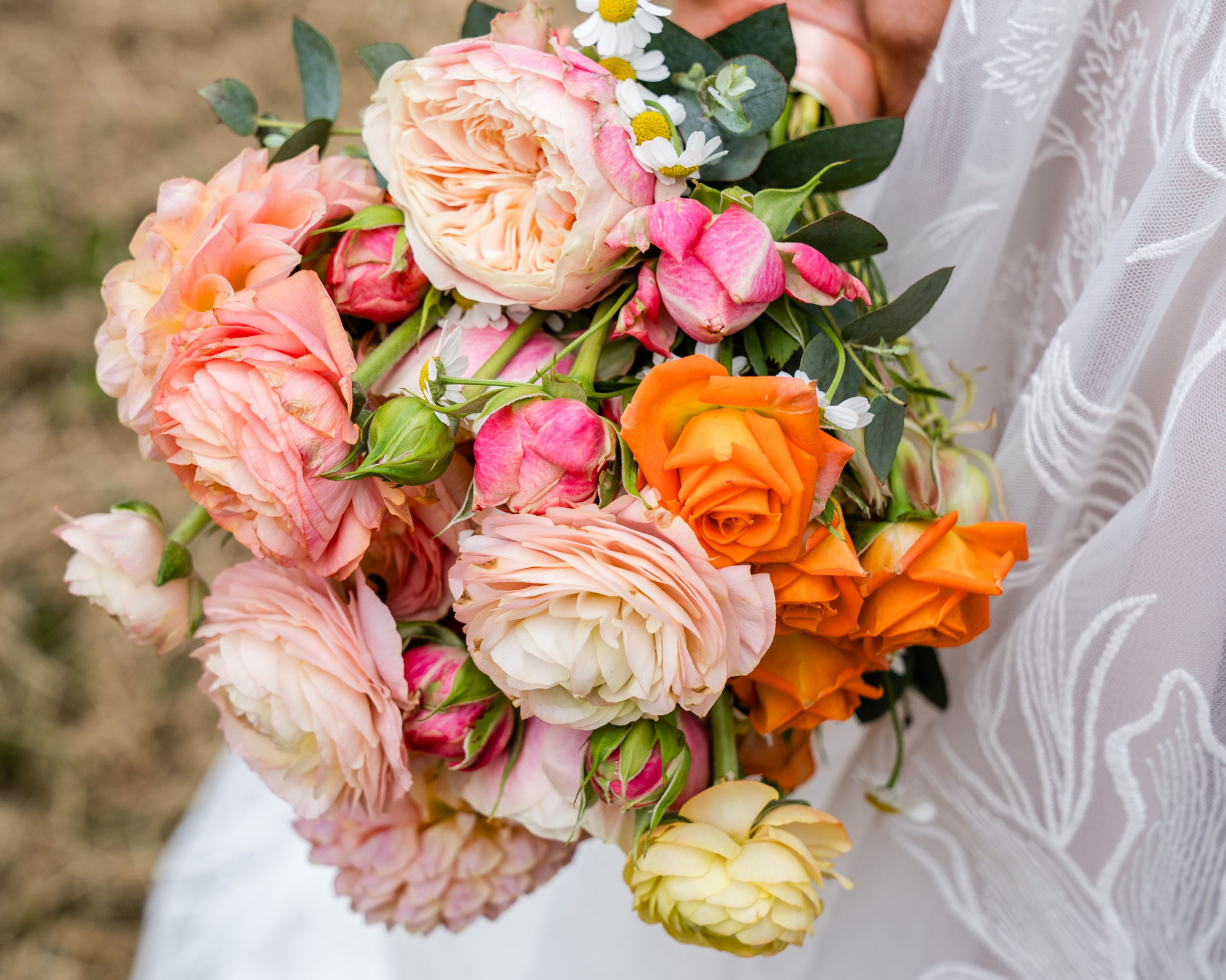Een bruidsboeket met roze, oranje en gele bloemen, gecombineerd met groen bladeren, vastgehouden door een bruid.