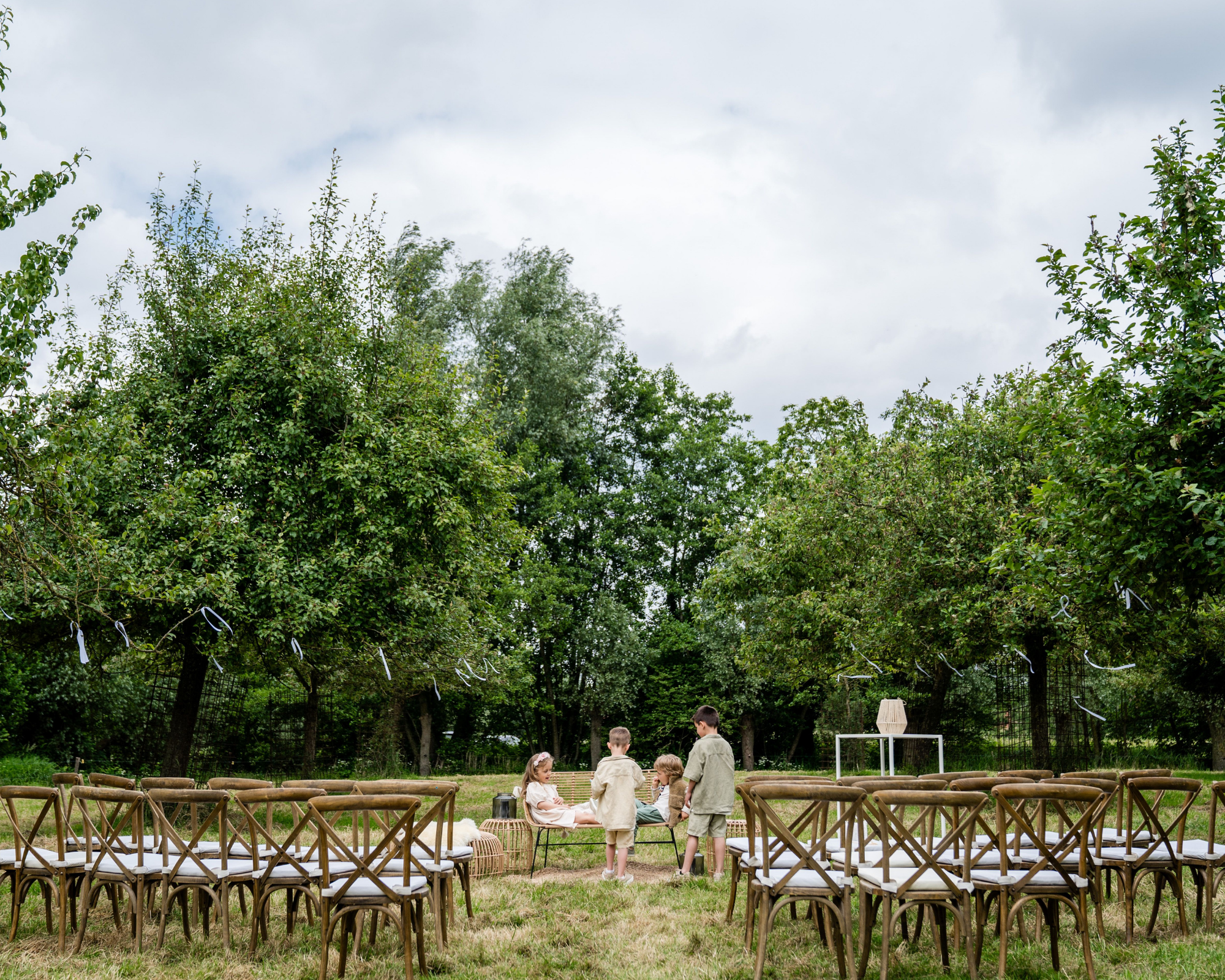 Kinderen spelen in een boomgaard met houten stoelen en groene bomen, klaar voor een ceremonie of buitenfeest.