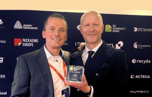 Deux hommes en costume posant pour une photo, l'un d'eux tenant un trophée ou un plaque lors d'une cérémonie ou d'une remise de prix.