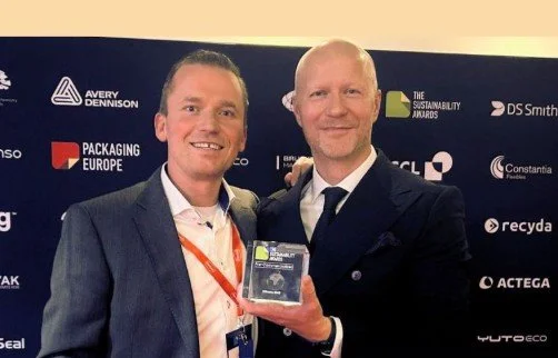 Deux hommes en costume posent pour une photo lors d'une cérémonie, l'un d'eux tient un trophée ou une plaque.