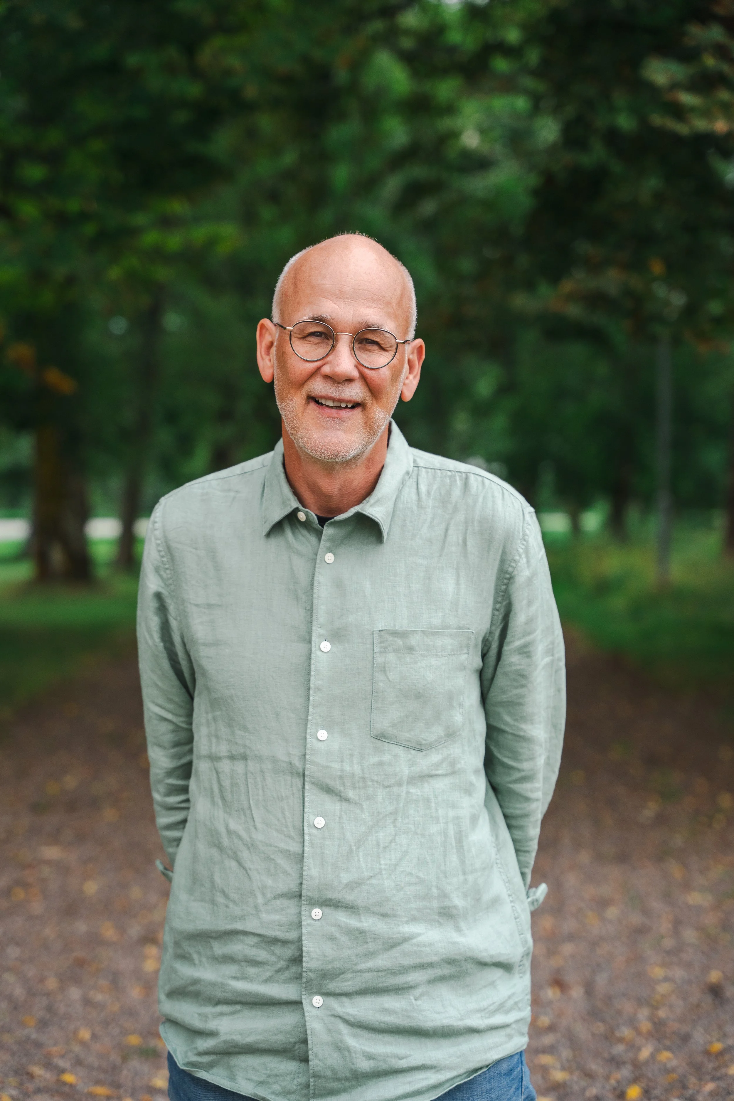 En äldre man med glasögon, grått skägg, blå jeans och en ljusgrön skjorta står på en skogsstig, omgiven av gröna träd och löv.