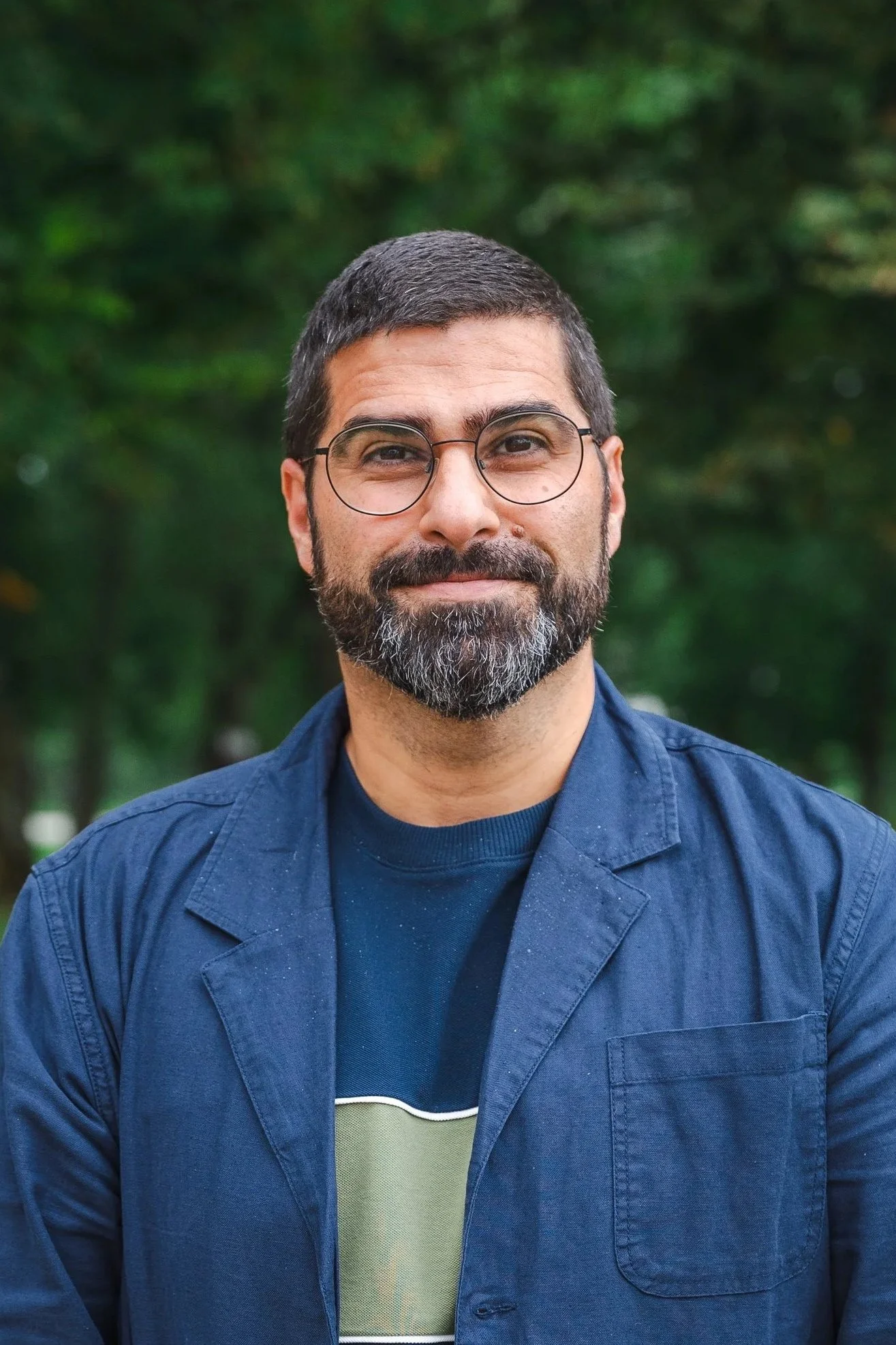 En man med mörkbrunt hår och skägg, bär glasögon och en blå jacka, står utomhus med en grön skog i bakgrunden.
