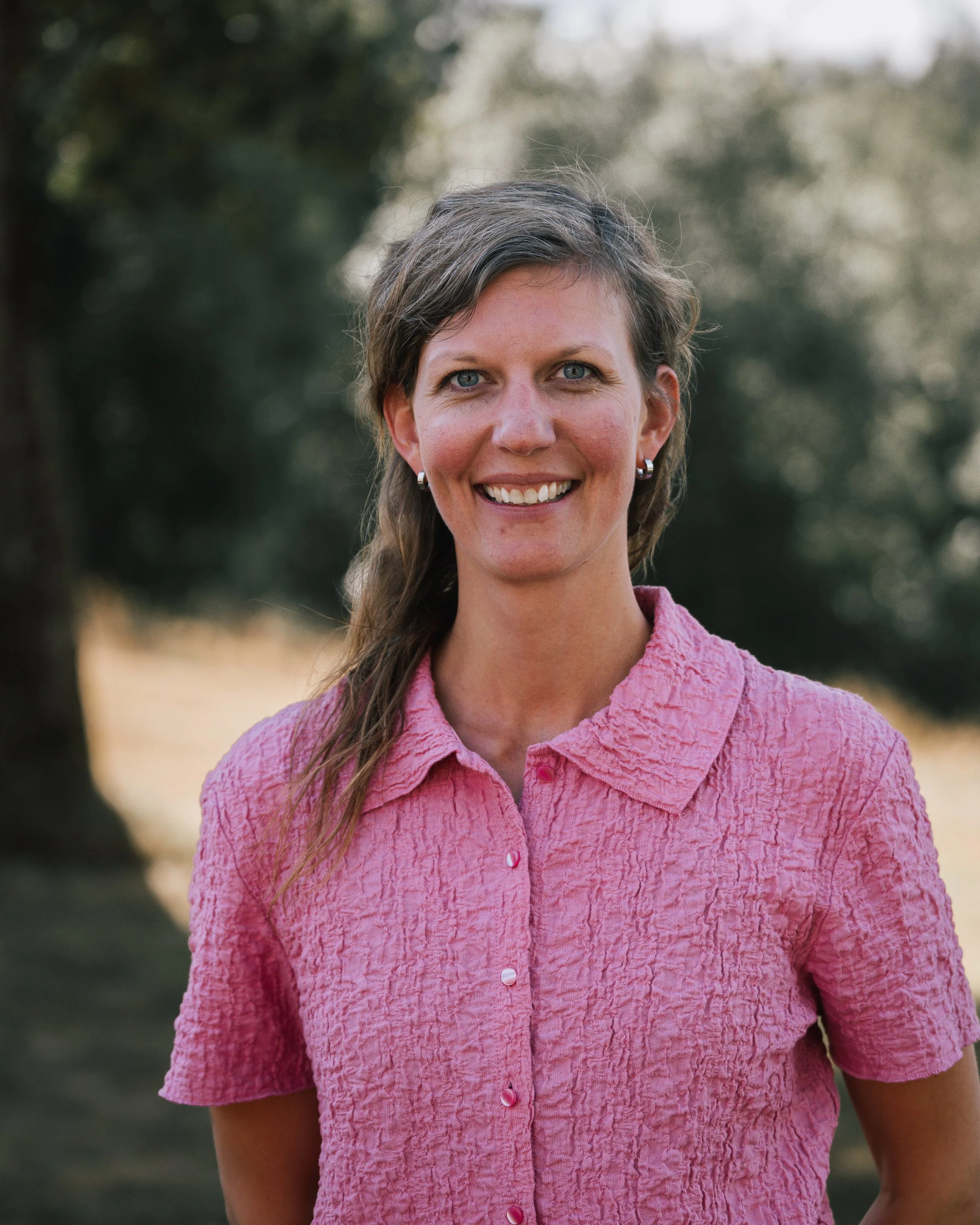 En woman med brunt, lockigt hår och ljusblå ögon, som ler, bär en rosa, knäppt skjorta och står utomhus med träd i bakgrunden.