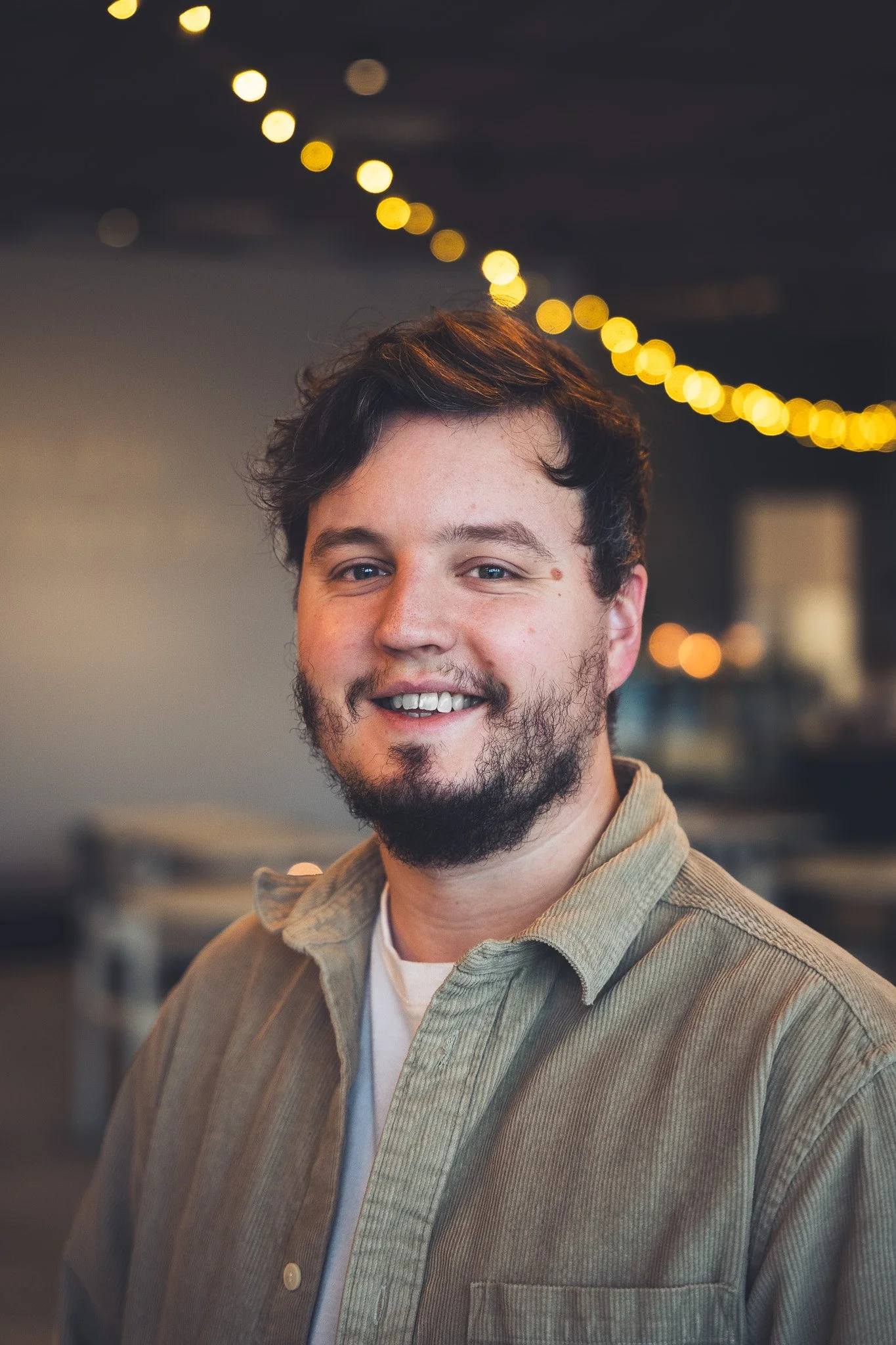 Ung man med mörkt, vågigt hår och skägg som ler, iklädd beige skjorta, mot en bakgrund av bokeh-lampor i ett inomhusmiljö.