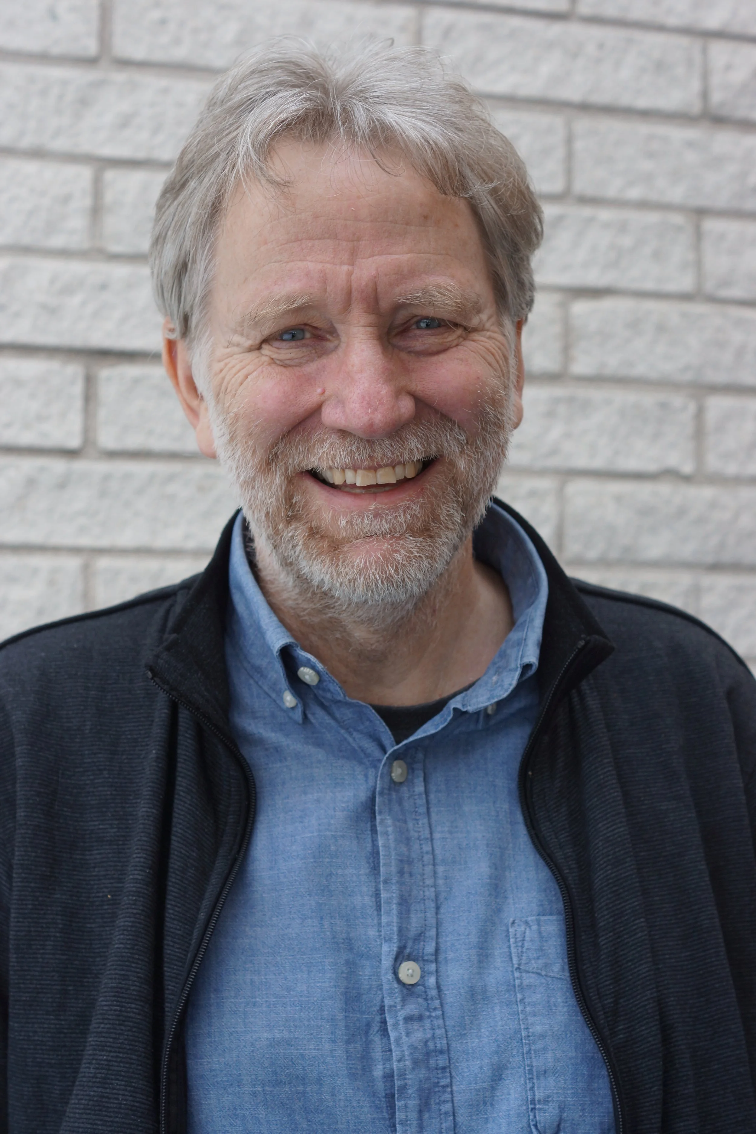 En äldre man med grått hår och skägg ler mot kameran, står framför en vit tegelvägg.