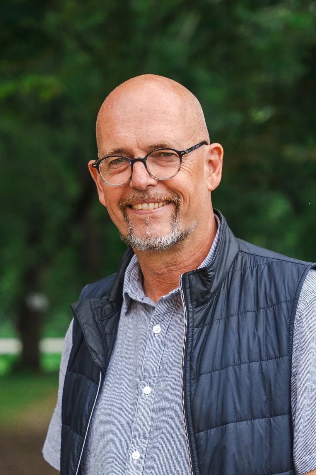 En leende man med glasögon, skägg och mustasch utomhus.