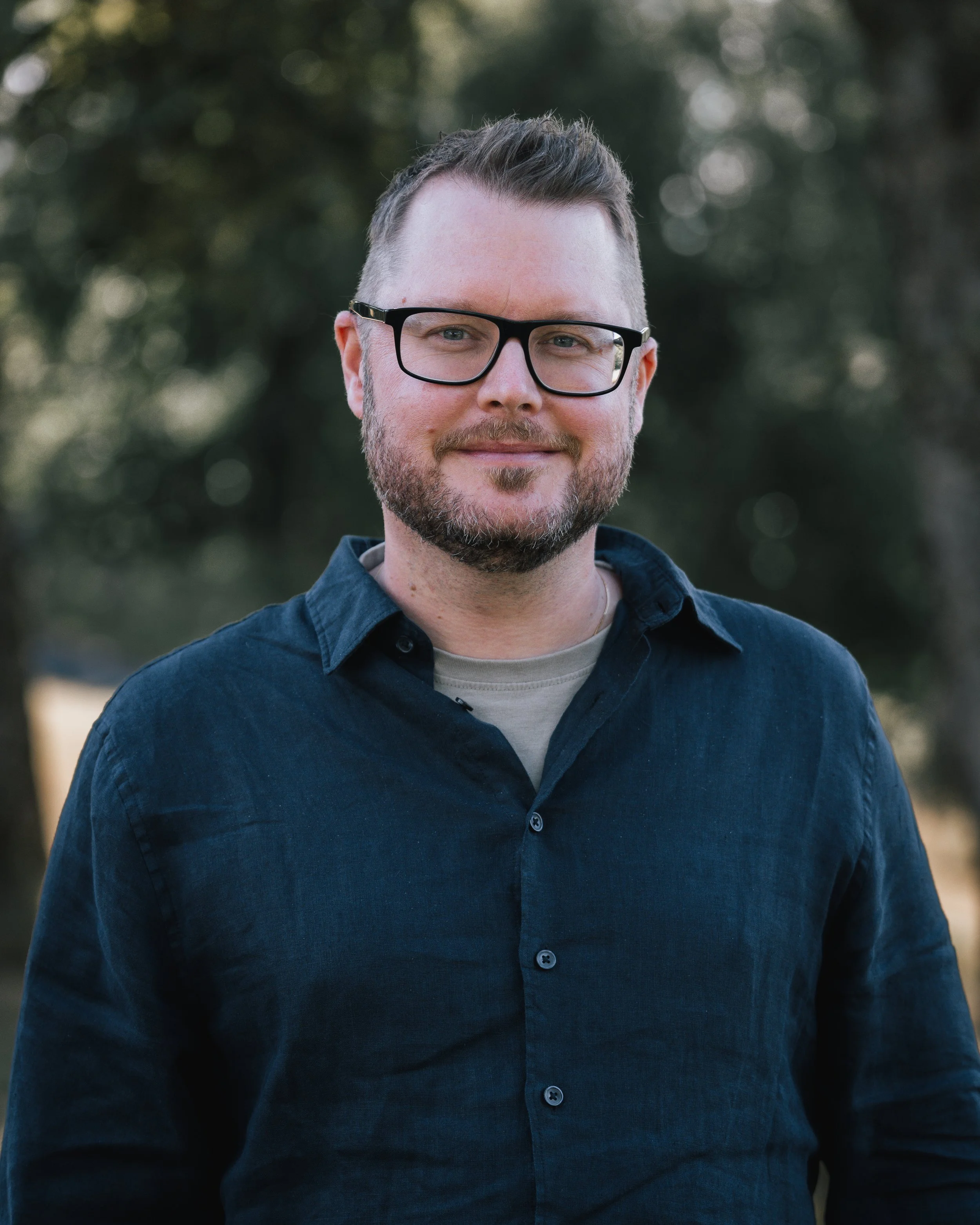 Porträtt av en man med glasögon, skägg och kort hår, utomhus med träd i bakgrunden.