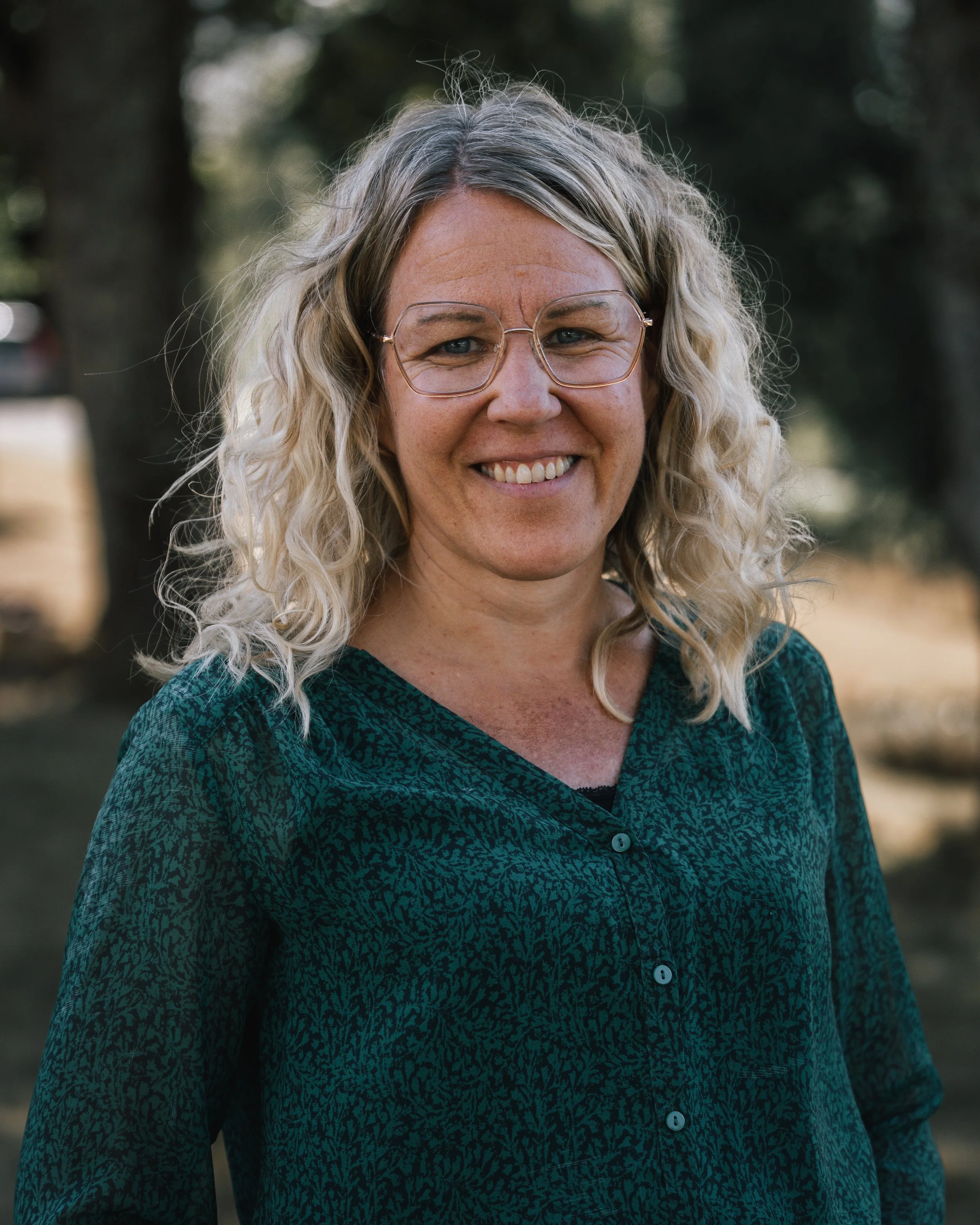 En leende kvinna med blont lockigt hår och glasögon, utomhus med träd i bakgrunden.