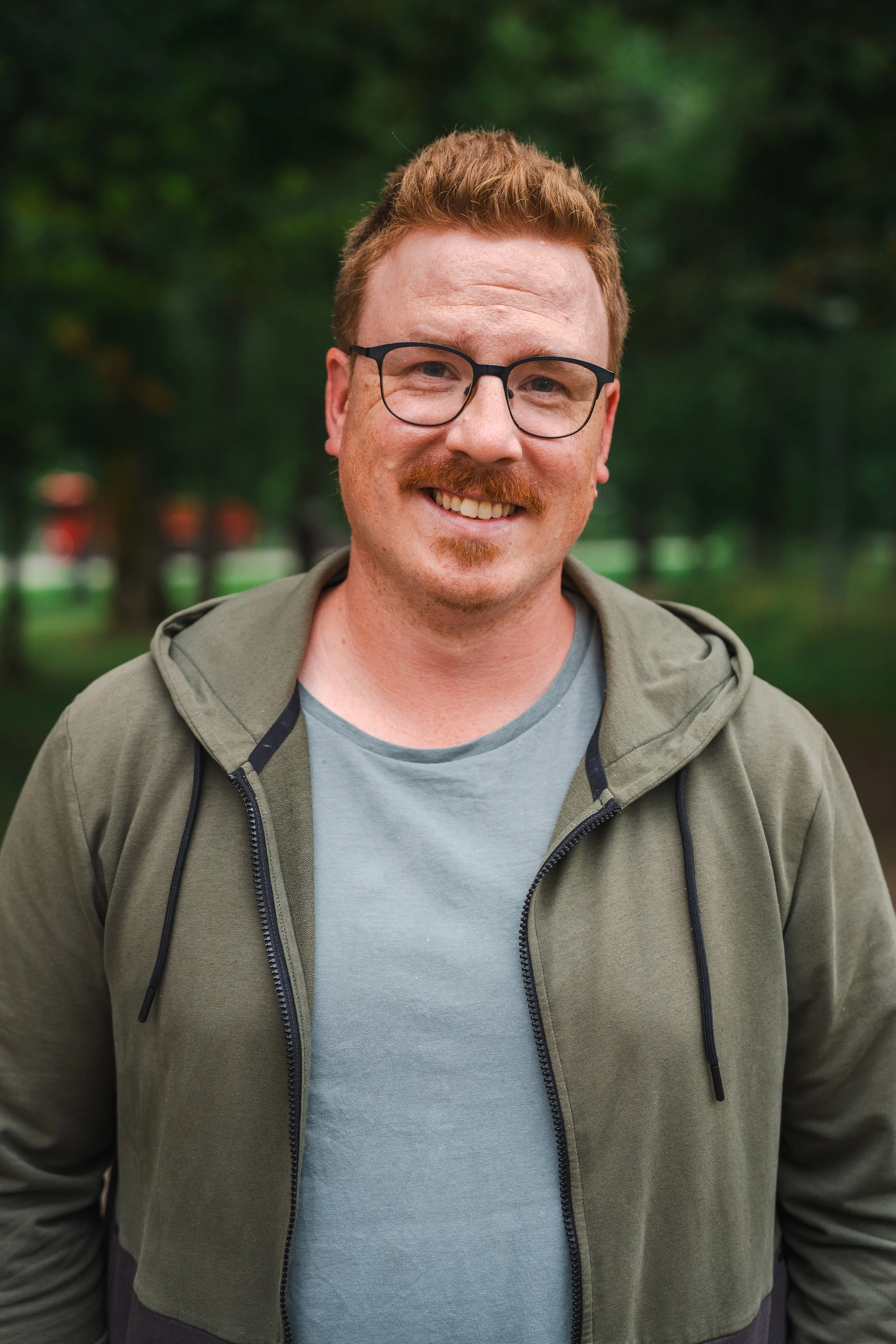 En man med rött hår, glasögon och skägg, ler och står utomhus i ett grönt parklandskap.