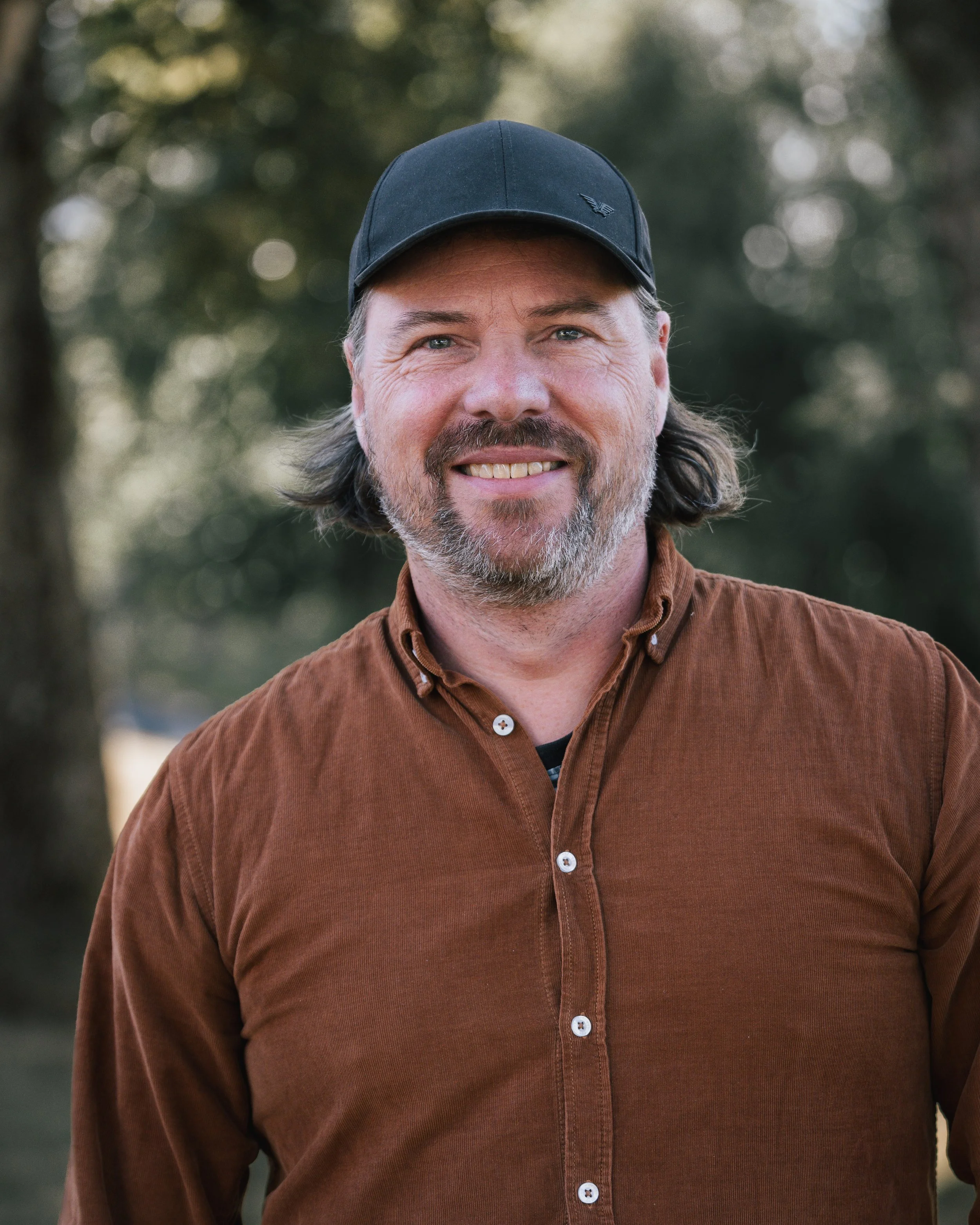 En man med grått hår och skägg, bär en svart keps och en brun skjorta, står utomhus i ett naturligt landskap.