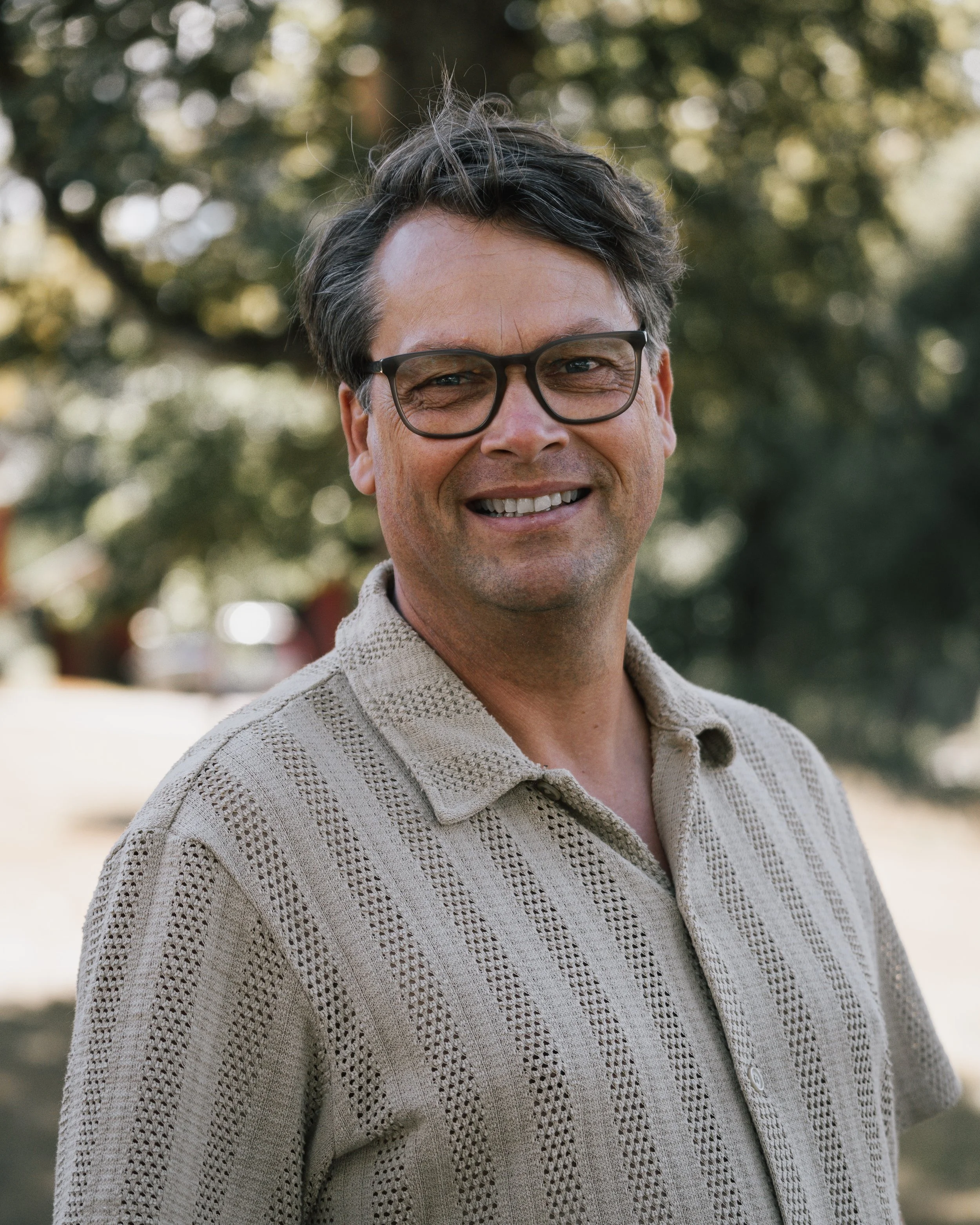 En man med mörkt hår och glasögon, ler, utomhus med träd i bakgrunden.
