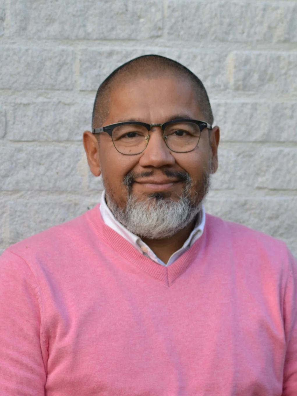 Man med glasögon och grått skägg, bär rosa tröja, står framför en ljus tegelvägg.