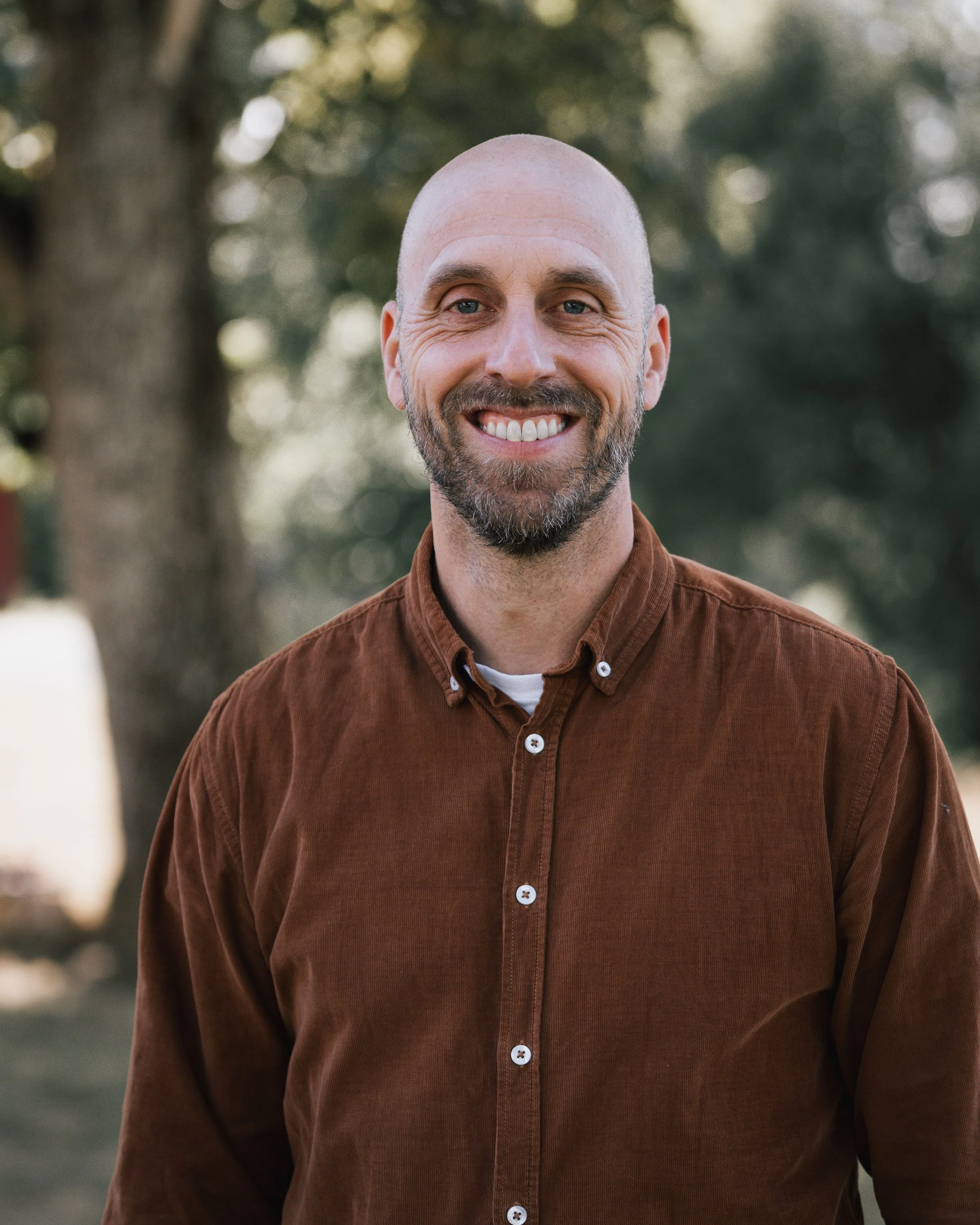 En man med skägg och skalle, iklädd brunt skjorta, står utomhus framför ett träd.
