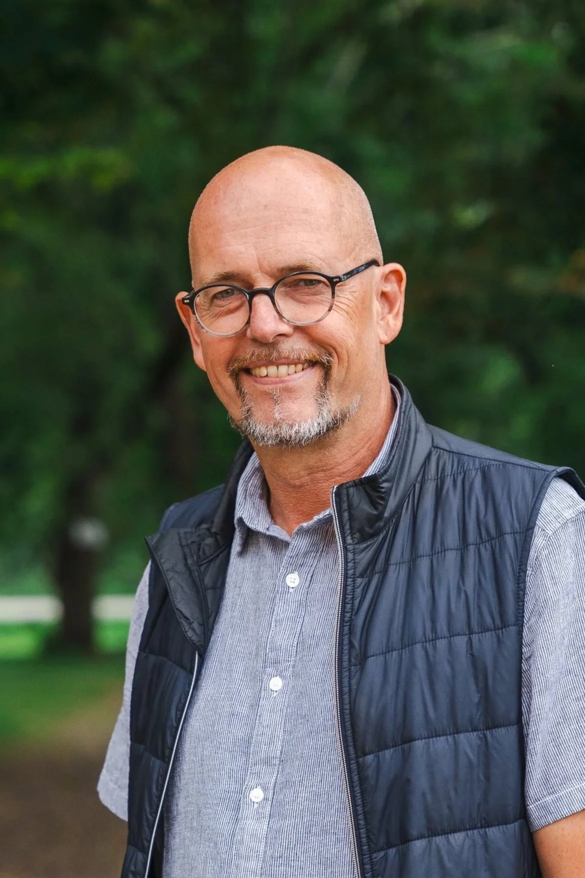 En leende man med glasögon och skägg, iklädd en grå skjorta och svart väst, står utomhus med gröna träd i bakgrunden.