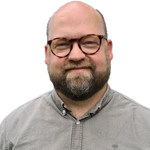 En man med glatt lockigt hår och glasögon, bär en grå skjorta.