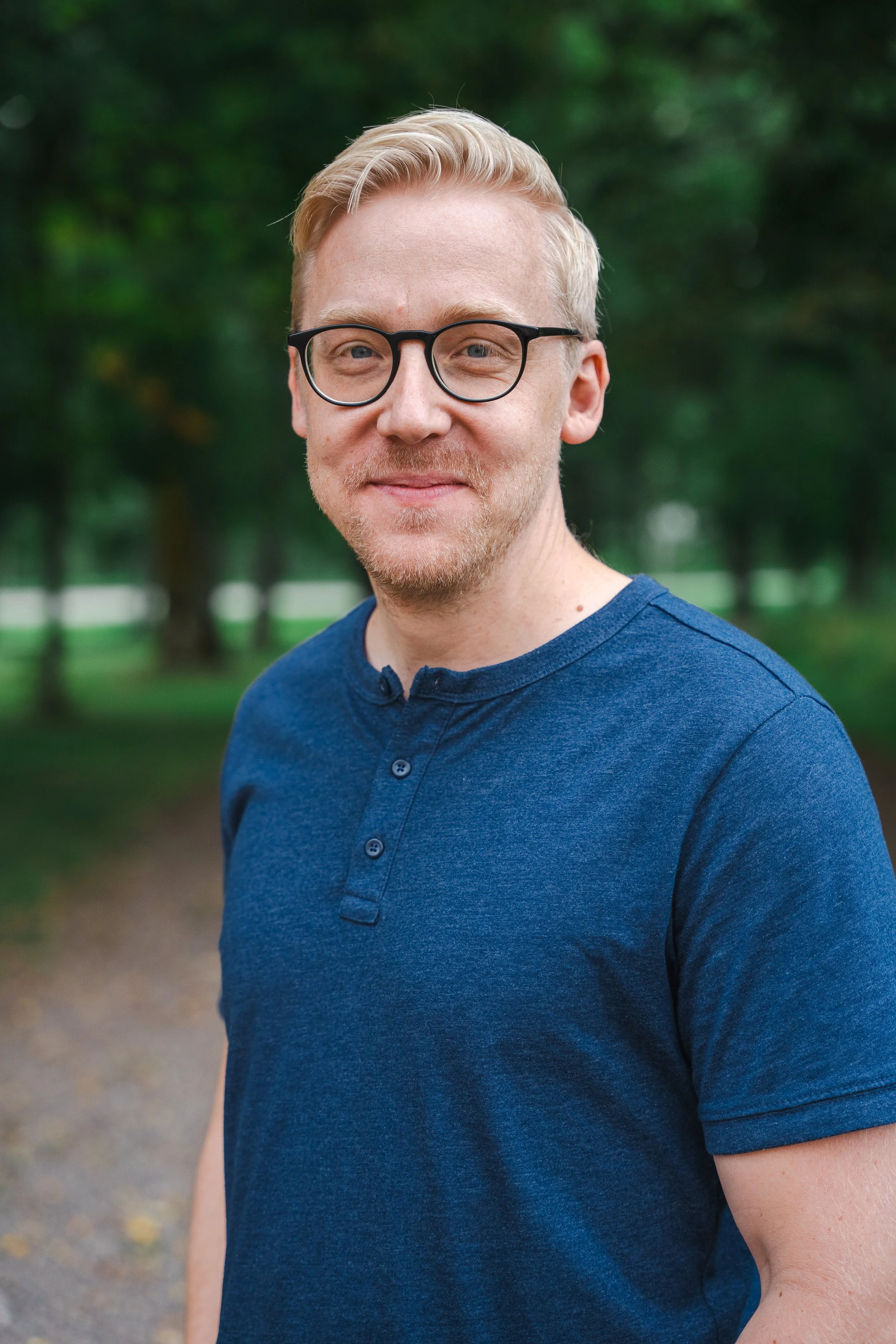 Porträtt av en man med glasögon, ljusblont hår, skägg, blå t-shirt, utomhus med gröna träd i bakgrunden.