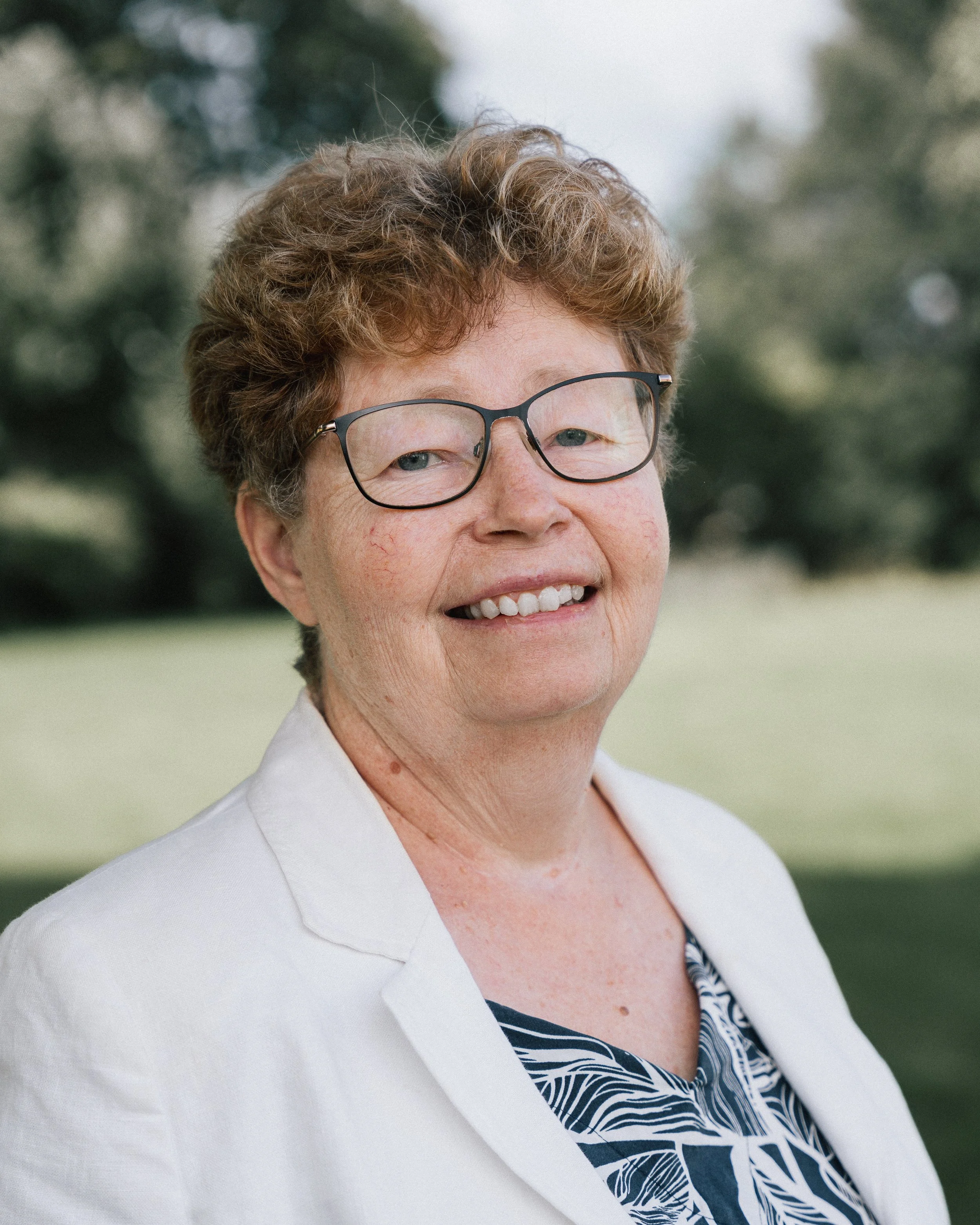 En äldre kvinna med kort, lockigt hår och glasögon, bär en vit blazer och blus med blå mönster, står utomhus med träd i bakgrunden.