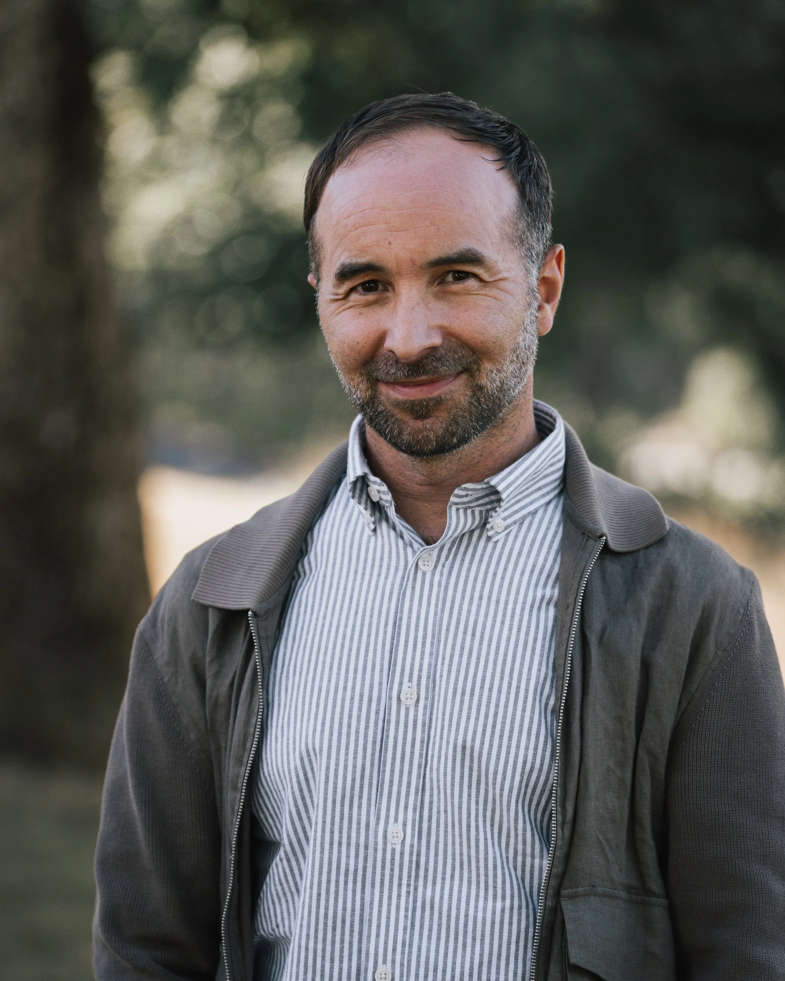 Ung man med kort mörkt hår och skägg ler, bär en randig skjorta och en grå jacka, står utomhus med träd i bakgrunden.