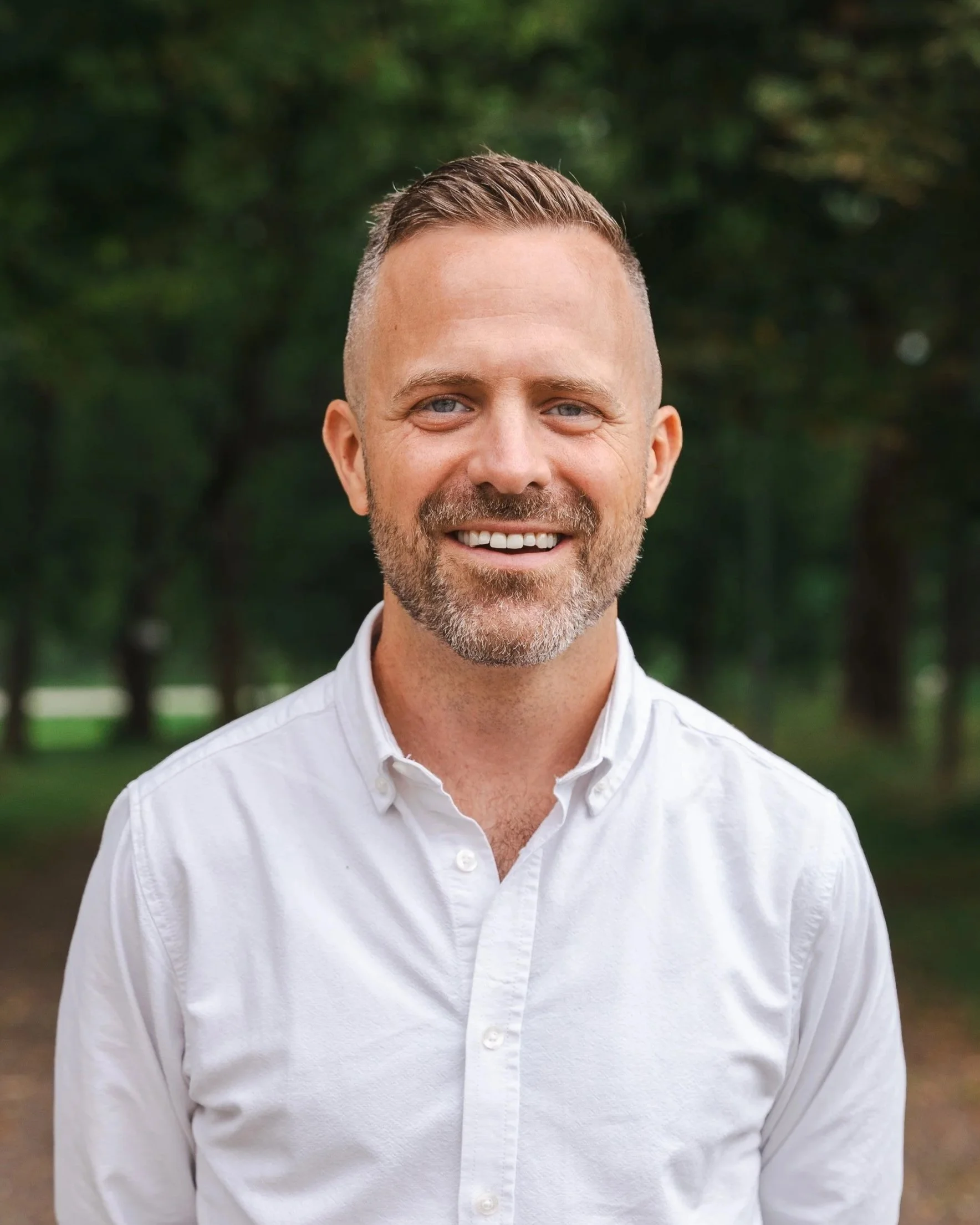 En man med ljus hud, kort tillbakatid, skägg, vitt skjorta, står utomhus i en park med gröna träd i bakgrunden och ler.