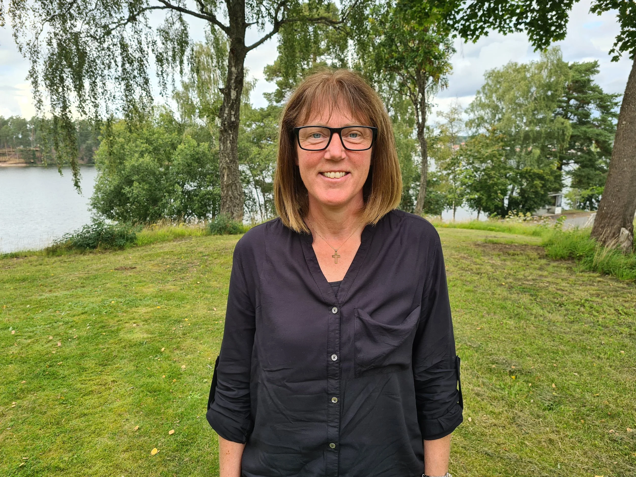 En kvinna med glasögon, brunt hår och svart skjorta står utomhus framför träd och en sjö, ler och tittar mot kameran.