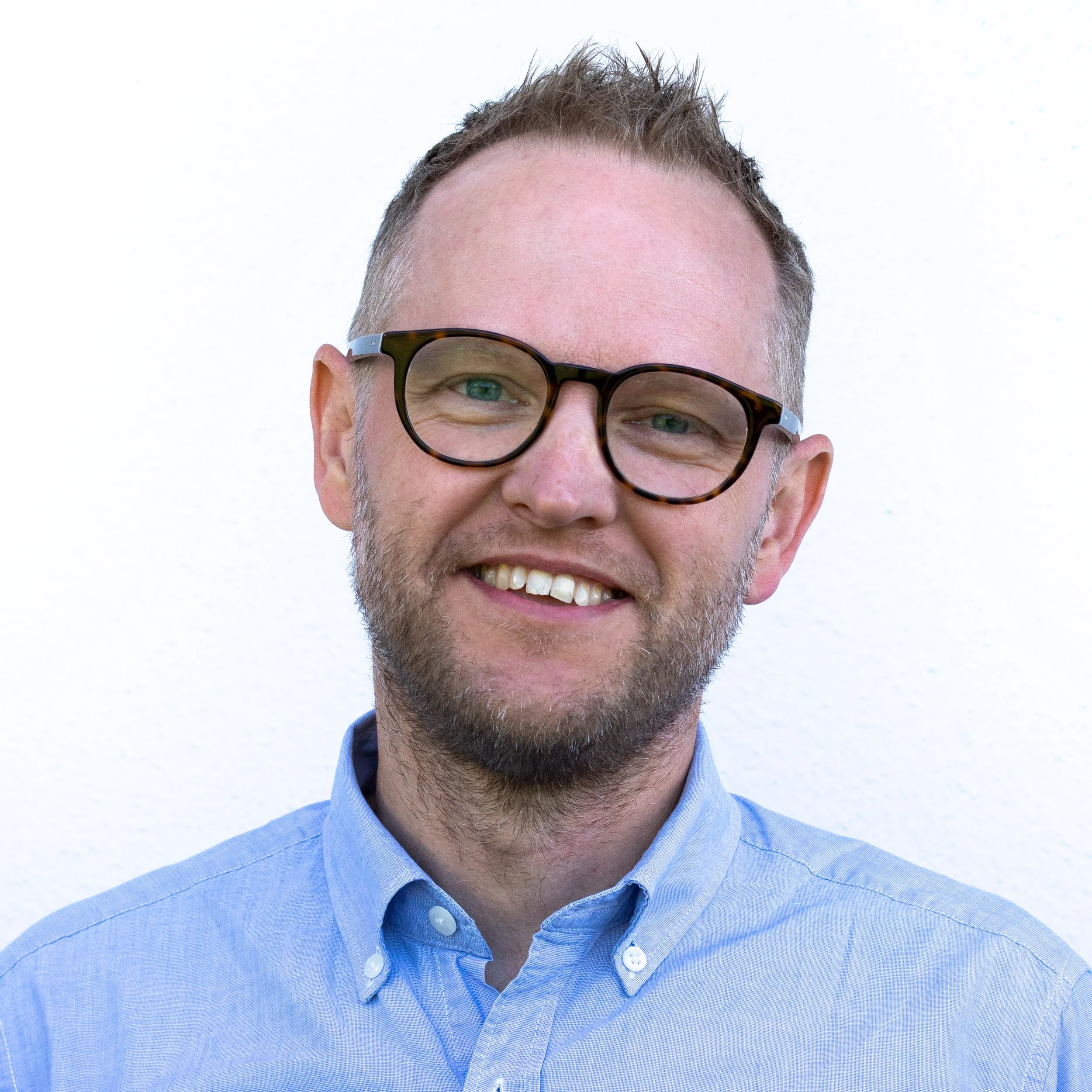 En man med glasögon och skägg ler mot kameran, bär en ljusblå skjorta, står framför en vit vägg.