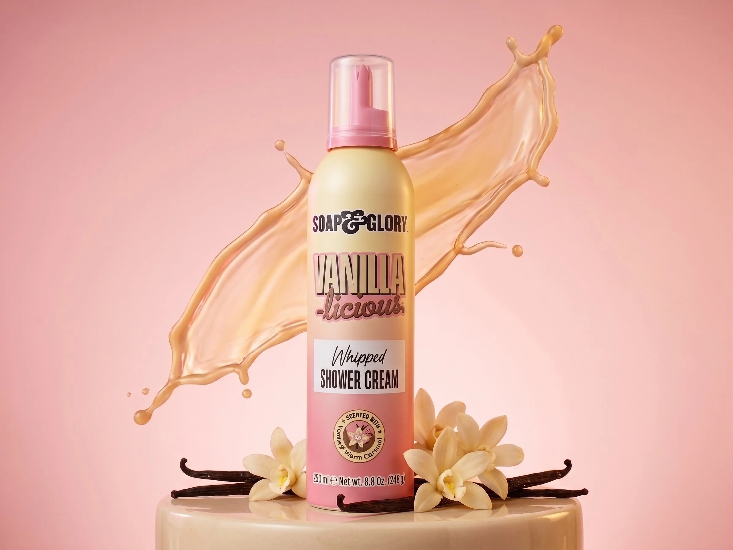 A pink shower foam bottle labeled 'Vanilla Licious' with vanilla flower and vanilla beans surrounding it, with vanilla and caramel-colored splashes in the background.