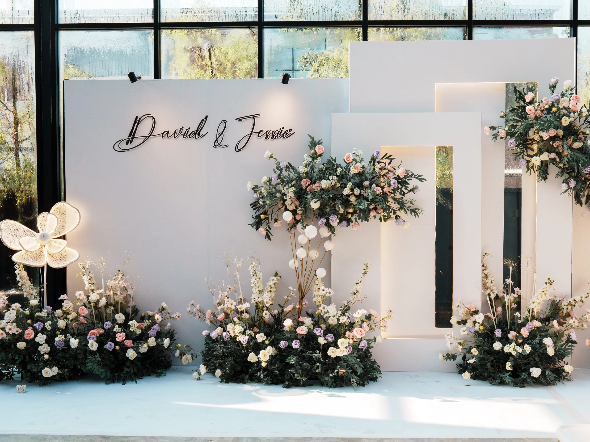 Wedding backdrop with white panels, floral arrangements, and a sign that says 'David & Jessie' in cursive black font.