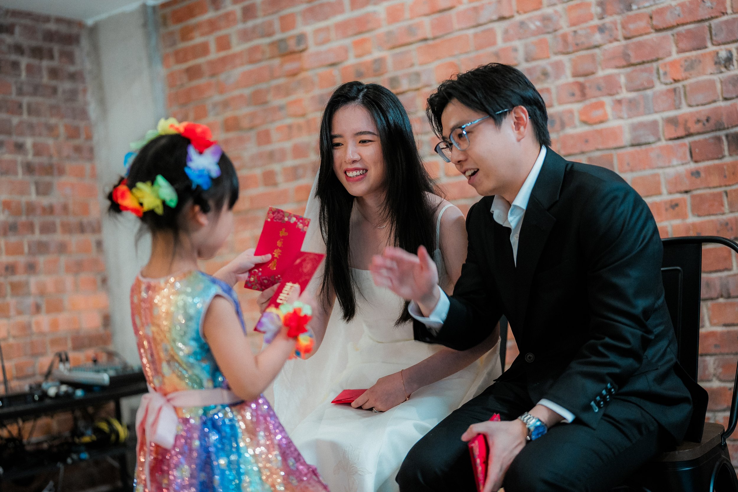 A young girl with colorful hair accessories in a glittery dress giving a red envelope to a smiling woman in a white dress and a man in a black suit, sitting against a brick wall.