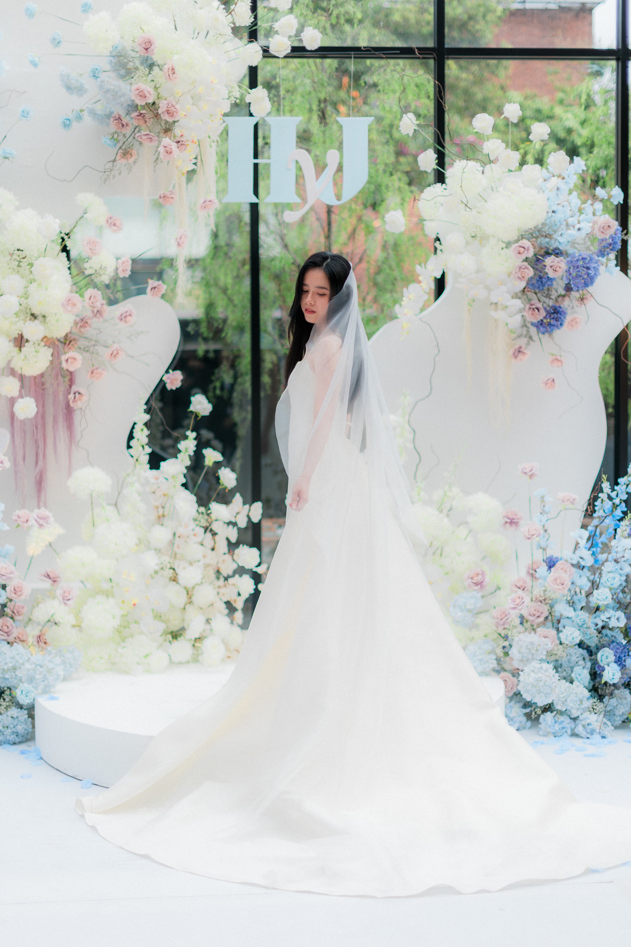 A woman in a white wedding gown and veil stands in front of a floral wedding decor with pink, white, and blue flowers, and large white backdrop pieces, with a window showing greenery in the background.