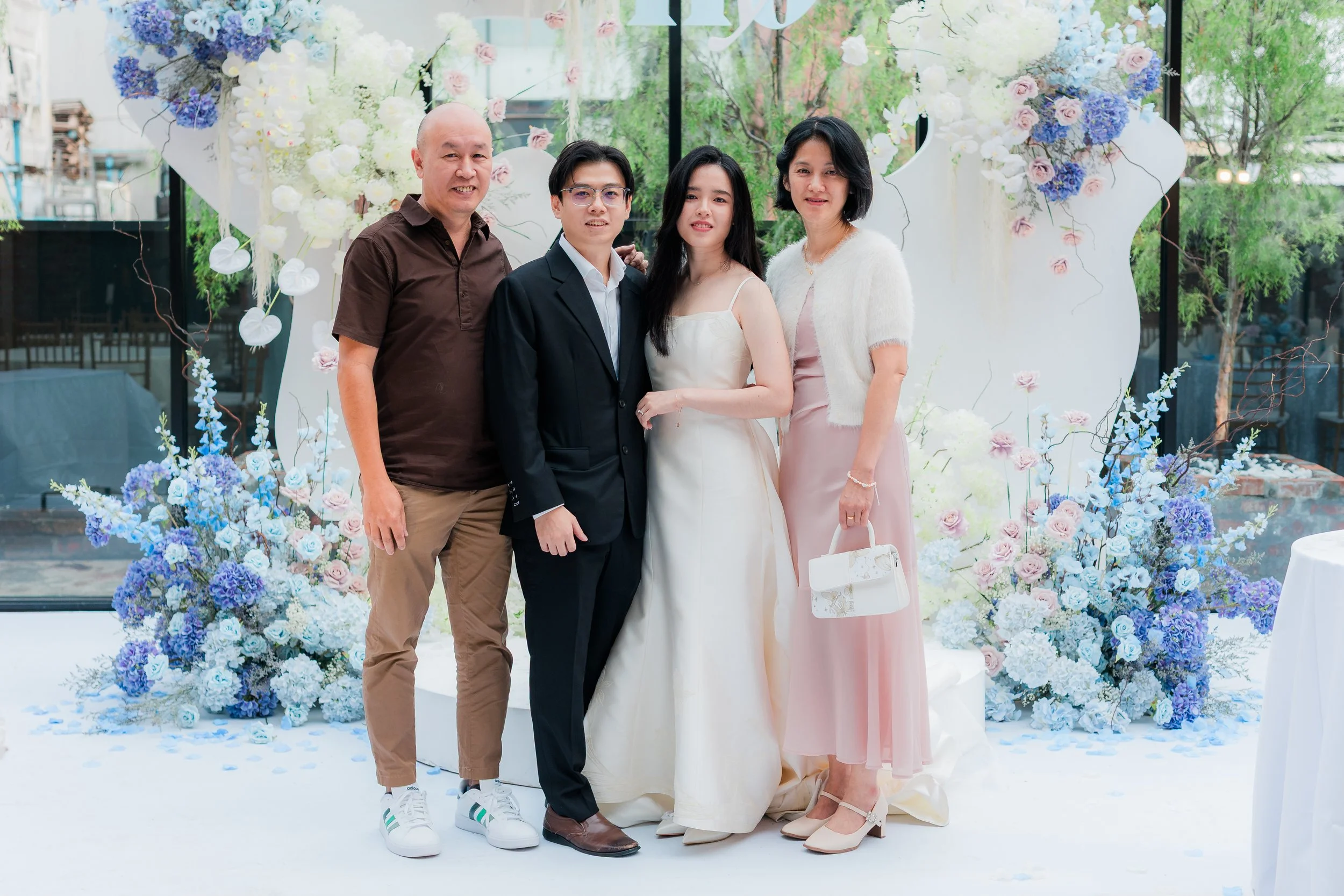 Four people standing in front of a floral wedding backdrop, including a bride in a white gown, a groom in a black suit, and two others, a man in a brown shirt and tan pants and a woman in a pink dress with a white cardigan, holding a white purse.